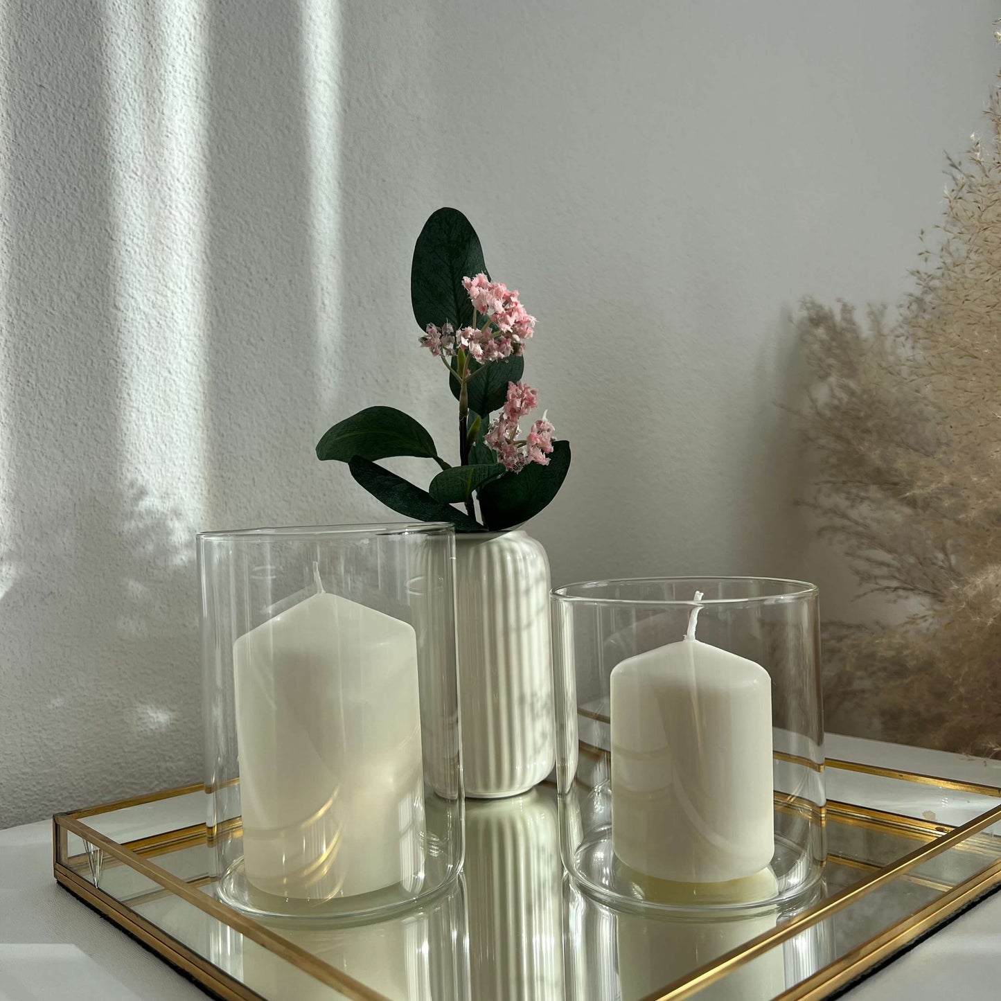 Zwei große Windlichtgläser mit weißen Kerzen auf einem goldgerahmten Spiegeltablett, ergänzt durch eine dekorative weiße Vase mit rosa Blüten – stilvolle und minimalistische Tischdekoration