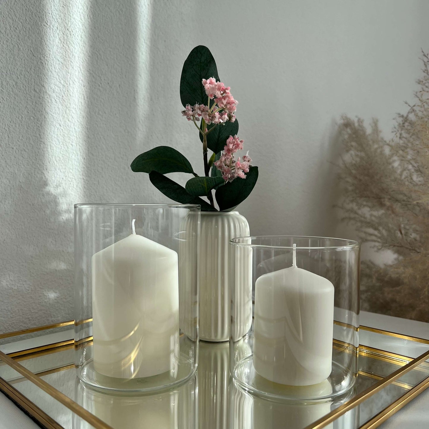 Zwei große Windlichtgläser mit weißen Kerzen auf einem goldgerahmten Spiegeltablett, ergänzt durch eine dekorative weiße Vase mit rosa Blüten – stilvolle und minimalistische Tischdekoration