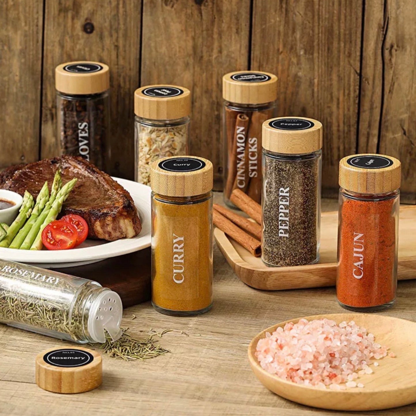 Eine rustikale Tischszene mit einer Auswahl an Gewürzstreuern, etikettiert mit ‘Curry’, ‘Pfeffer’, ‘Cajun’ und anderen. Sie sind auf einem Holztisch platziert neben einem Teller mit einem saftigen Steak, Spargel und Tomaten, was die Verwendung der Gewürze in der Küche zum Kochen und Servieren von Mahlzeiten unterstreicht.
