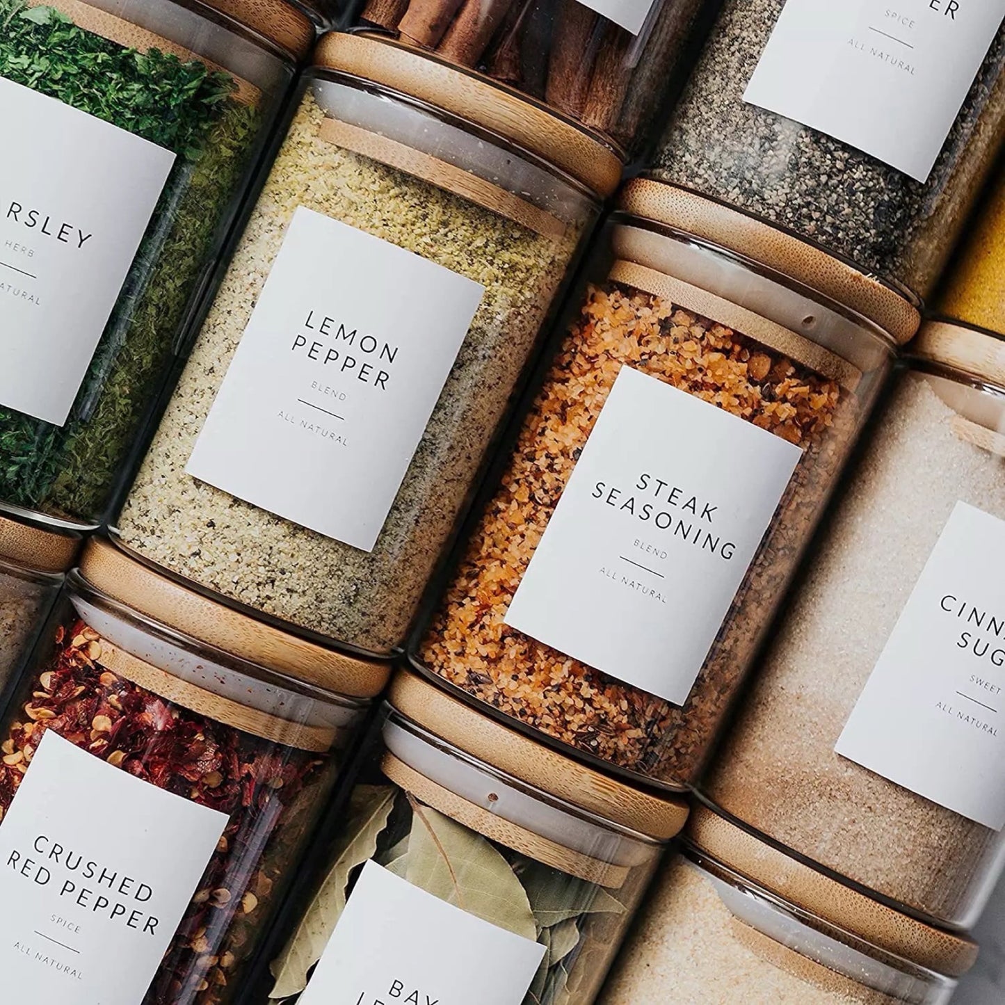 Eine Reihe von Gewürzgläsern mit Bambusdeckeln, ordentlich beschriftet mit verschiedenen Gewürzen wie Petersilie, Zitronenpfeffer und Steakgewürz. Die Gläser sind dicht nebeneinander aufgereiht, zeigen die vielfältigen Farben und Texturen der Gewürze und unterstreichen eine organisierte und stilvolle Küchenumgebung.