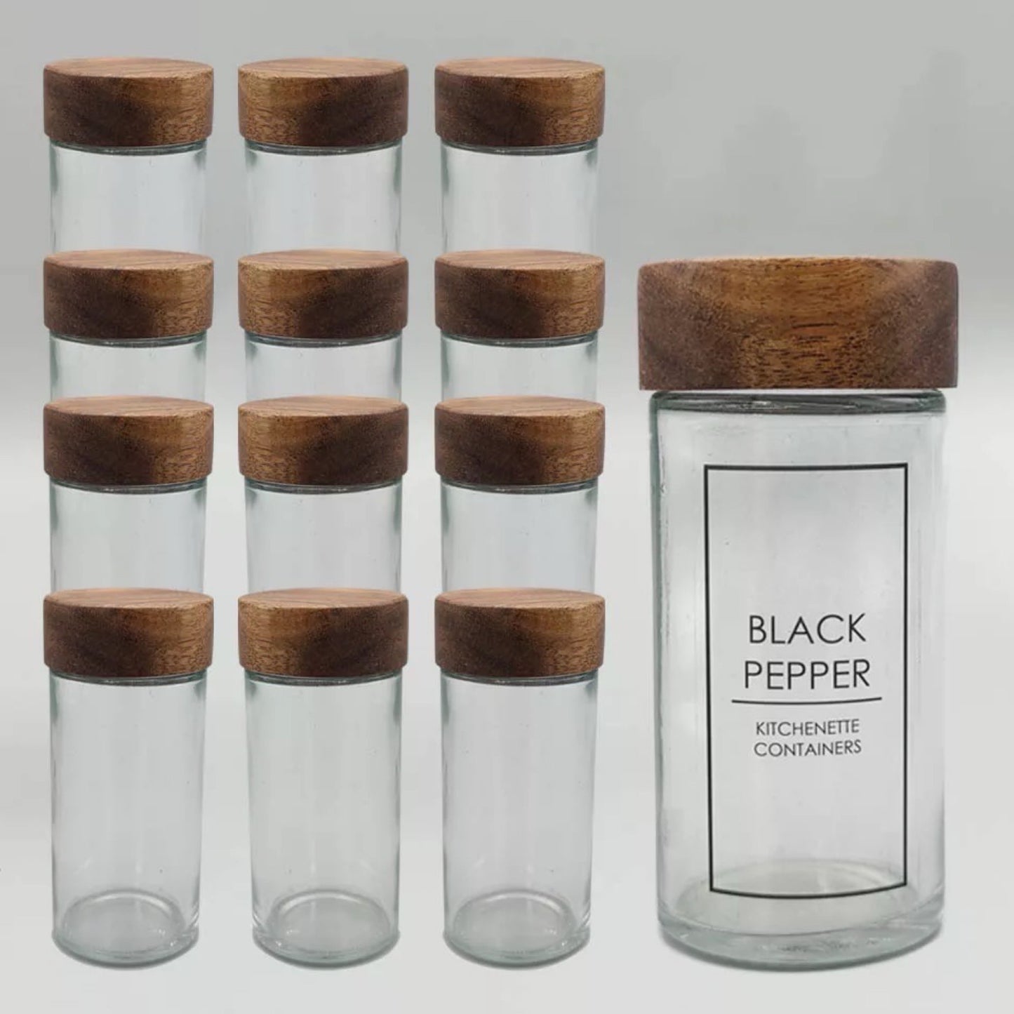 Eine Kollektion von 12 leeren, transparenten Gewürzgläser mit robusten Akazienholzdeckeln, ordentlich aufgereiht. Eines der Gläser ist mit einem Etikett versehen, das ‘BLACK PEPPER’ beschriftet ist, was die Verwendung dieser Behälter zur Aufbewahrung von Gewürzen in der Küche nahelegt.
