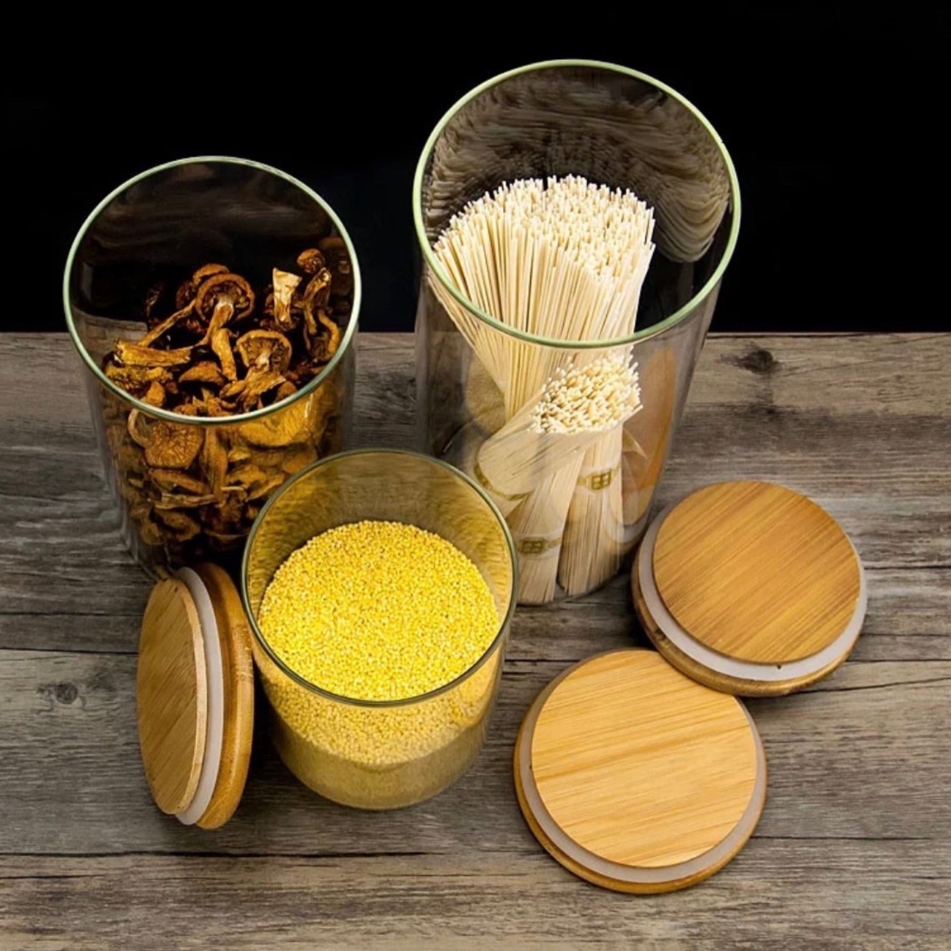 Set aus klaren Glasbehältern mit Bambusdeckeln auf einem rustikalen Holztisch. Die Gläser enthalten getrocknete Pilze, gelben Hirse, und Bündel von Holzspießen. Diese Aufbewahrungslösung bietet nicht nur einen Blick auf die Inhalte, sondern auch eine ästhetische Präsentation von Küchenzutaten und Zubehör.