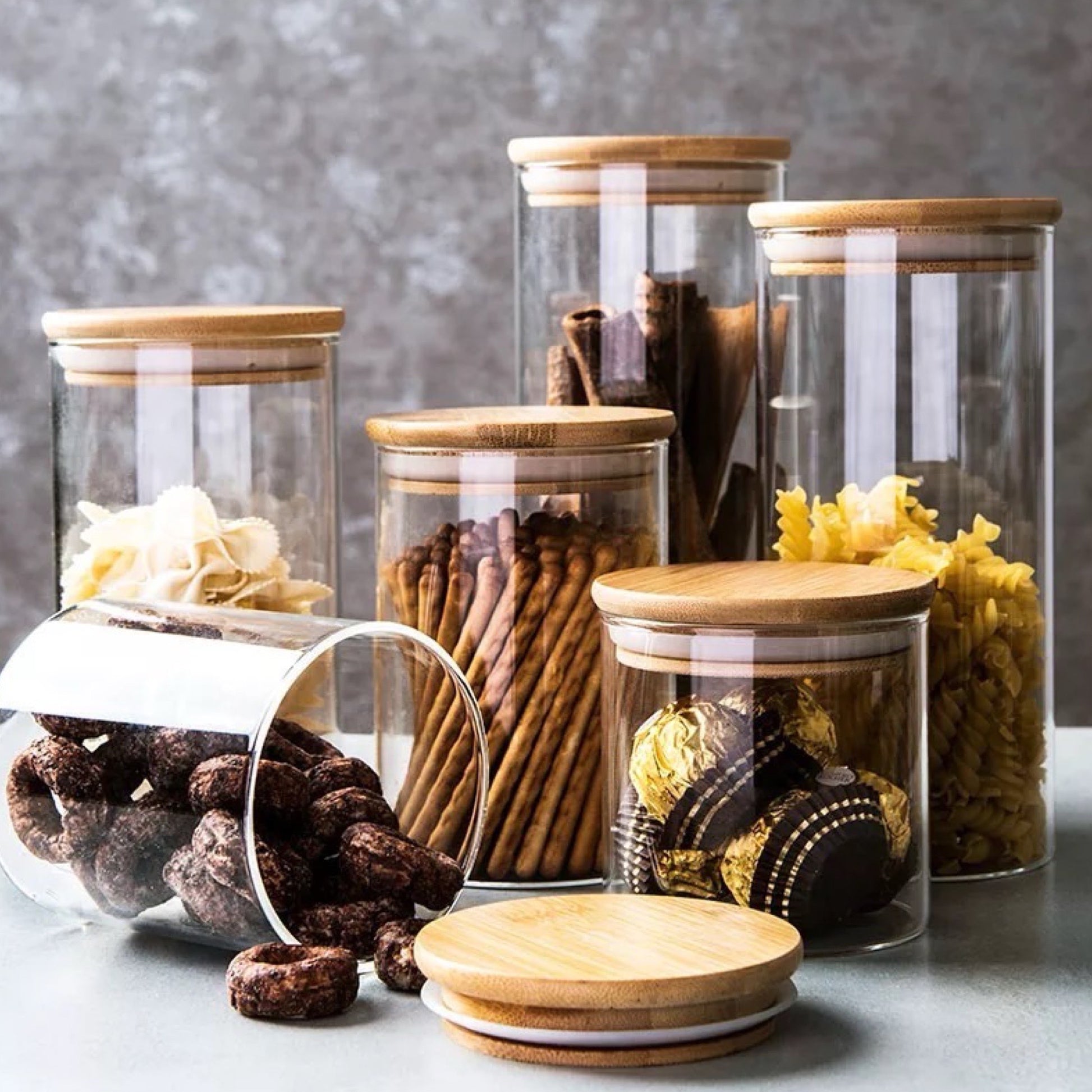 Vielseitige Kollektion von durchsichtigen Glasvorratsbehältern mit Bambusdeckeln auf einer grauen Unterlage. In den Gläsern befinden sich verschiedene Backzutaten und Snacks, darunter Schokoladenkekse, Brezeln, Fusilli-Nudeln und Zimtstangen. Ein Glas liegt auf der Seite und Schokoladenkekse sind daneben gestreut, was für eine einladende und praktische Aufbewahrungslösung spricht.