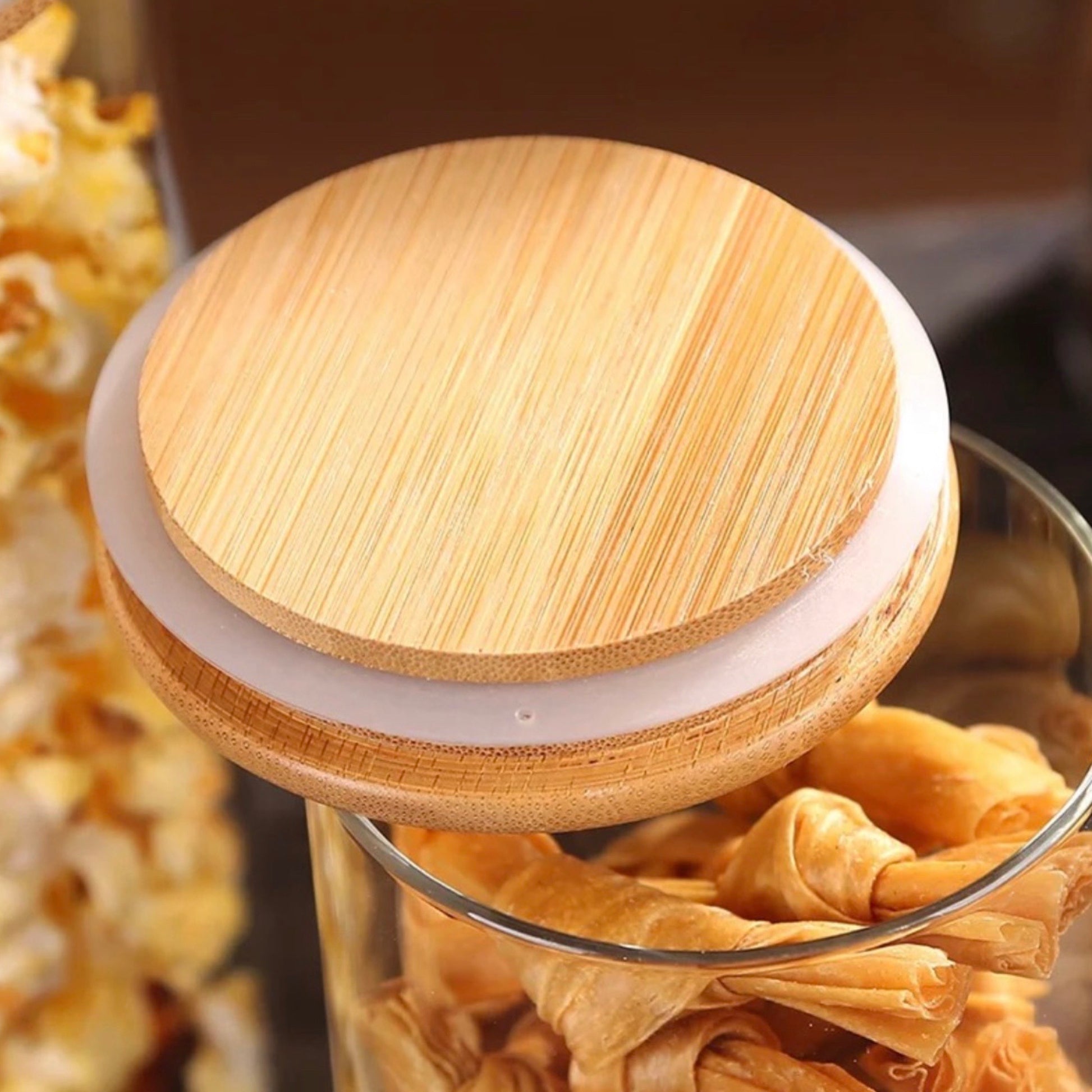 Nahaufnahme eines eleganten Vorratsglasdeckels aus Bambus mit einer glatten Oberfläche und feinen Maserungsdetails. Der Deckel sitzt auf einem klaren Glasbehälter, der mit knusprigen Blätterteigstückchen gefüllt ist, was eine stilvolle und funktionale Lösung zur Aufbewahrung von Snacks bietet.