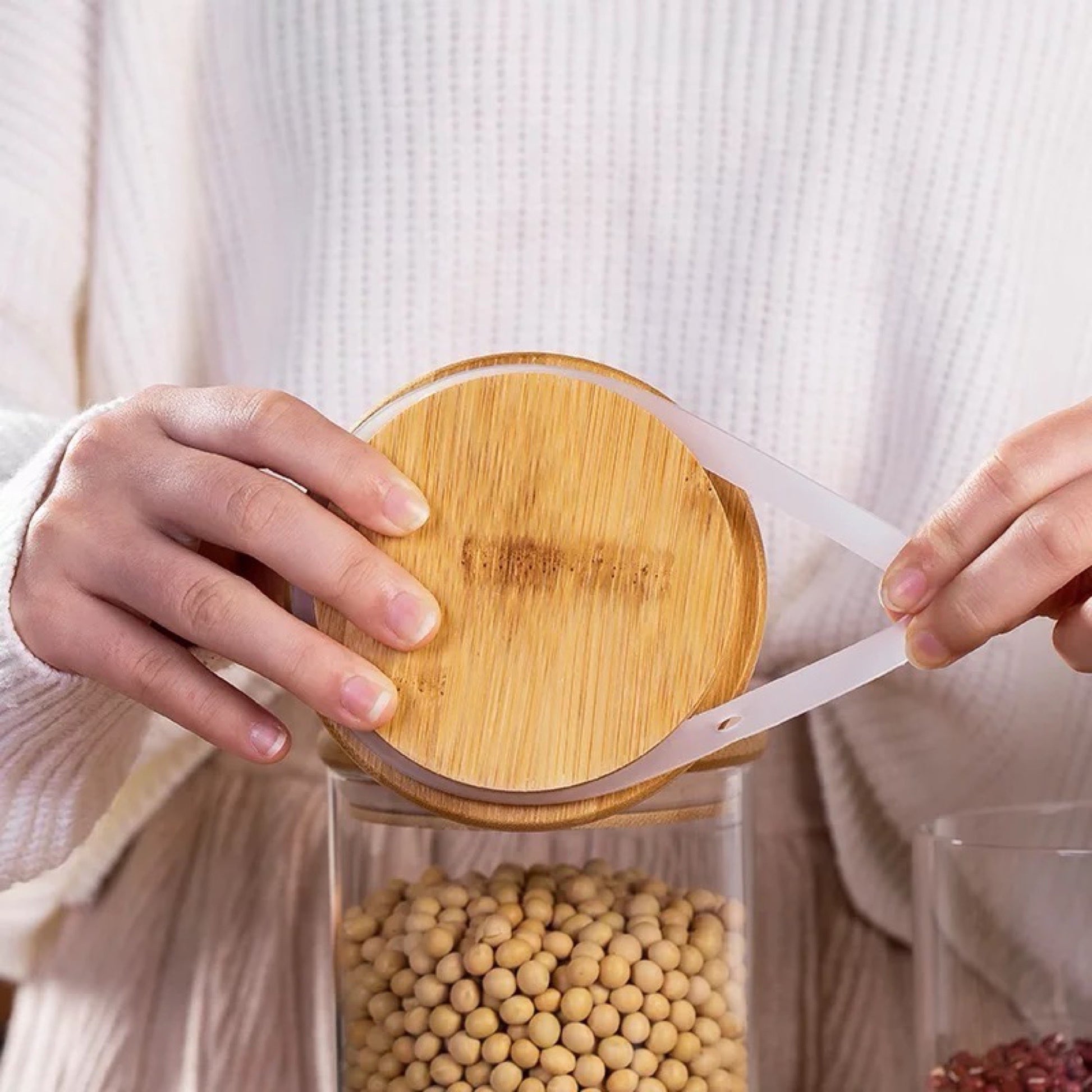 Nahaufnahme von Händen, die einen runden Bambusdeckel auf einem Glasbehälter platzieren. Das Glas ist gefüllt mit Sojabohnen, sichtbar hinter dem durchsichtigen Glas. Dieses Bild betont die Benutzerfreundlichkeit und natürliche Ästhetik der Vorratsgläser, ideal für die Aufbewahrung verschiedener Trockenvorräte in der Küche.