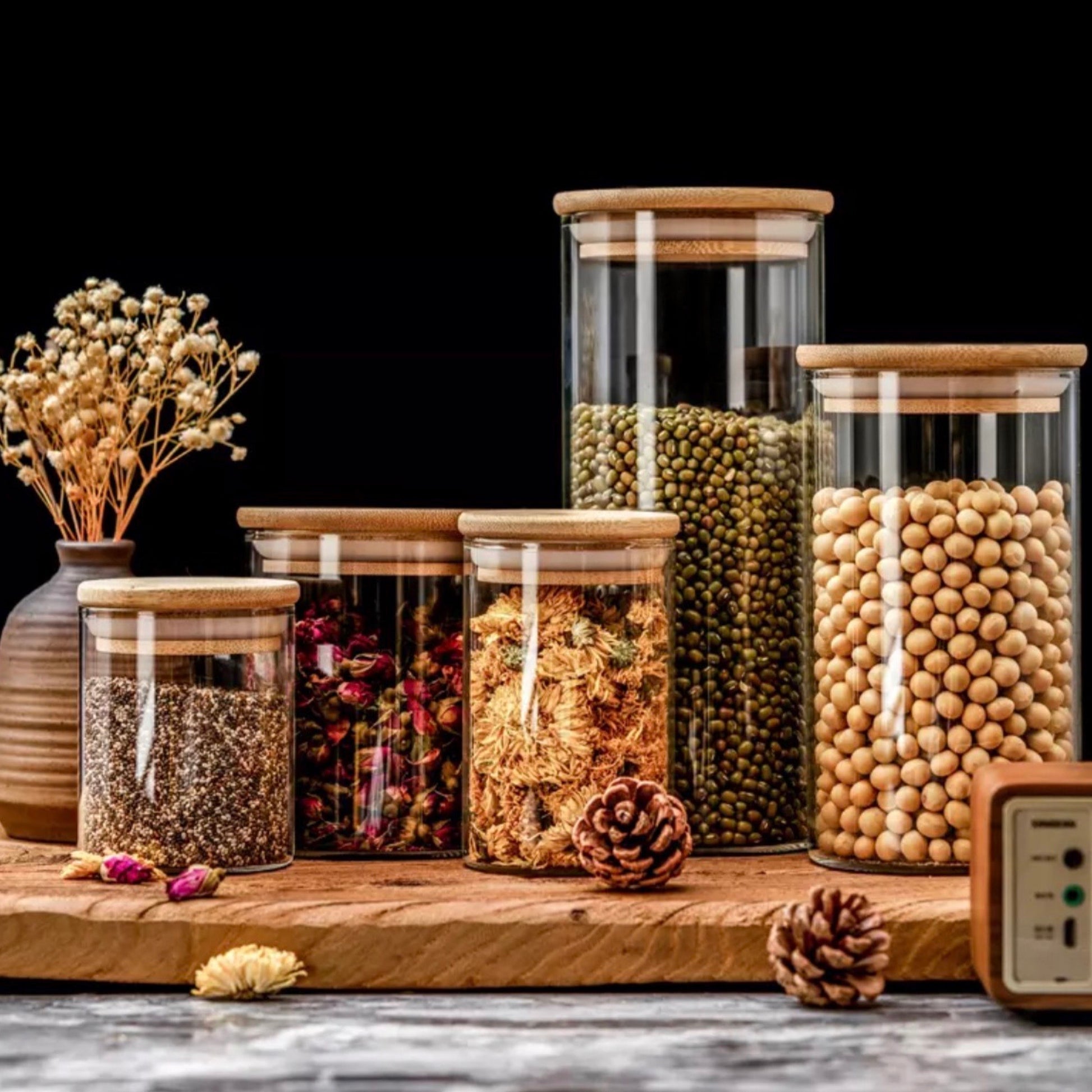 Stilvolle Küchenanordnung mit einer Reihe von transparenten Glasvorratsbehältern mit Bambusdeckeln, gefüllt mit einer Vielfalt von trockenen Lebensmitteln und Dekorationsobjekten. Zu sehen sind Gläser mit grünen Erbsen, Sojabohnen, Chiasamen, getrockneten Blumen und Kräutern, arrangiert auf einer Holzoberfläche neben einer rustikalen Vase mit Trockenblumen und Kiefernzapfen, was eine gemütliche und organische Ästhetik vermittelt.