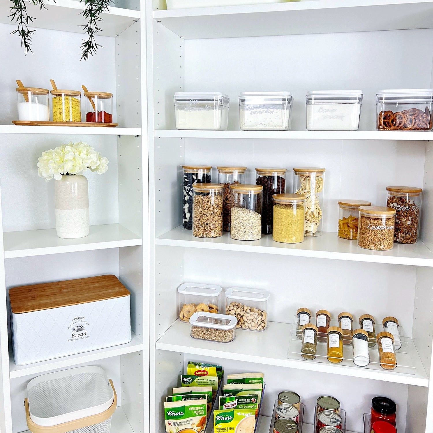Offenes weißes Küchenregal mit verschiedenen Vorratsdosen, kleinen Gläsern, einem Brotkasten und dekorativer Vase – perfekte Speisekammerorganisation mit praktischen Aufbewahrungslösungen.