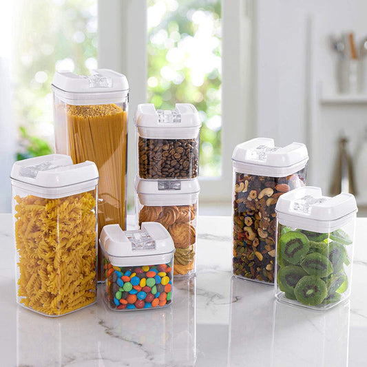 Verschiedene transparente Vorratsbehälter mit weißem Deckel auf einer hellen Küchenarbeitsplatte, gefüllt mit Lebensmitteln wie Spaghetti, Kaffeebohnen, Mandeln, gemischten Nüssen, getrockneten Kiwischeiben und bunten Süßigkeiten, beleuchtet durch natürliches Licht, das durch ein Fenster einfällt.