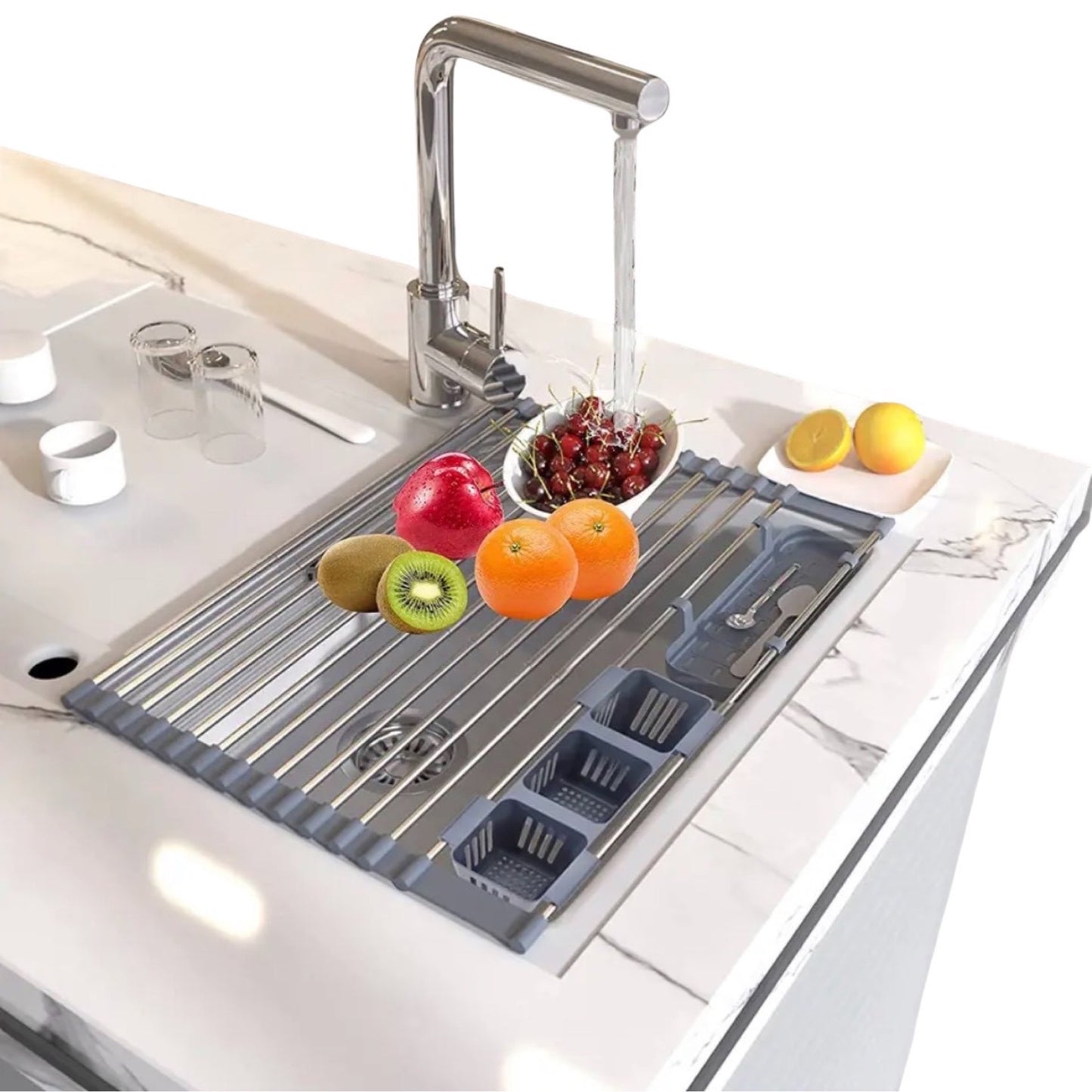 Modernes Abtropfgestell aus Edelstahl über einem Küchenspülbecken mit laufendem Wasserhahn, ausgestattet mit einem Besteckhalter und rutschfesten Silikonenden. Frische Früchte wie Kiwis, Orangen, und ein Granatapfel befinden sich auf dem Gestell neben einem Teller mit halbierter Zitrone und zwei Gläsern.