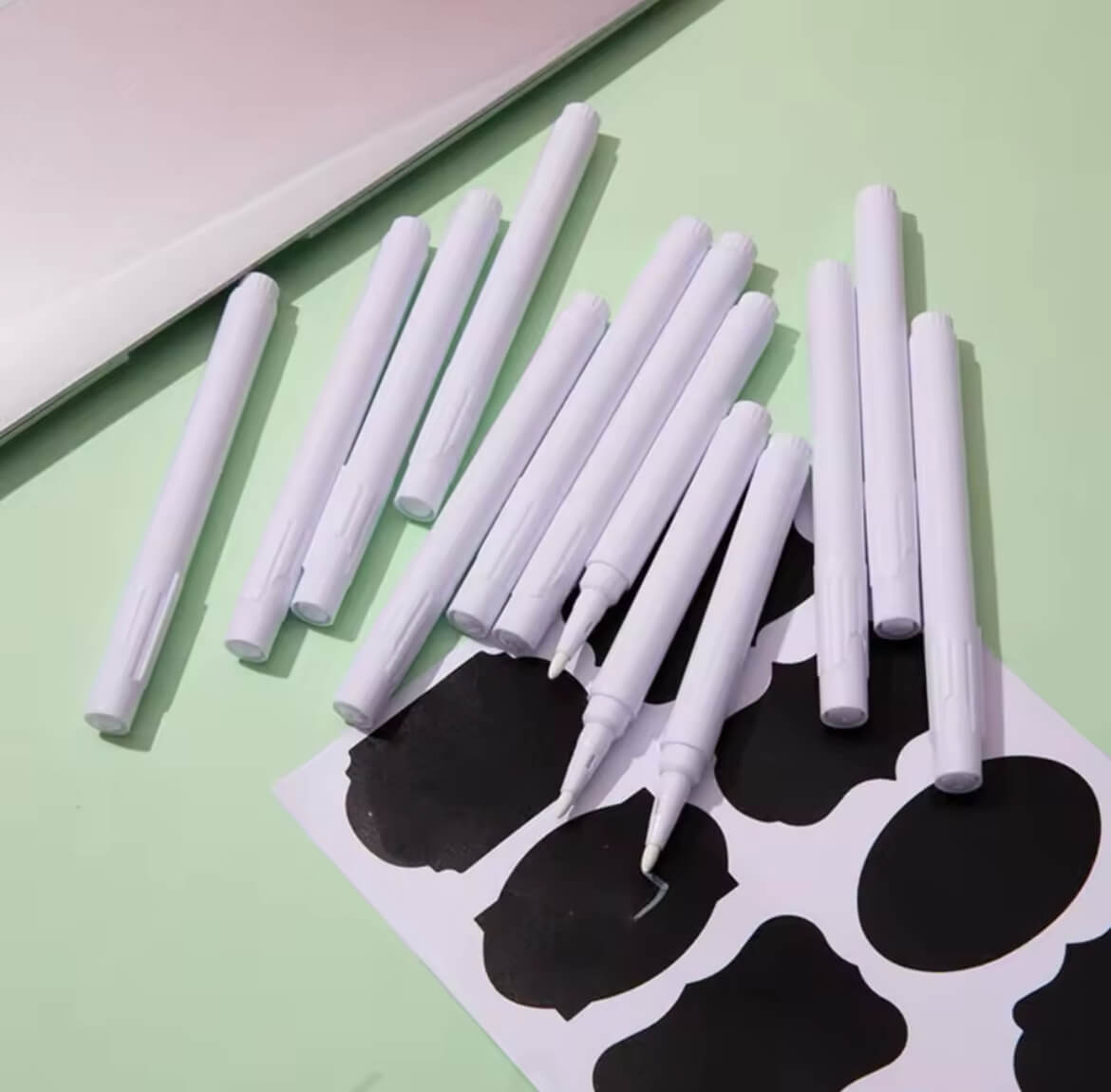 Set aus weißen Kreidemarker mit  schwarzen Etiketten, arrangiert auf grünem Hintergrund – ideal zum Beschriften von Vorratsdosen, Gläsern oder Aufbewahrungsboxen.