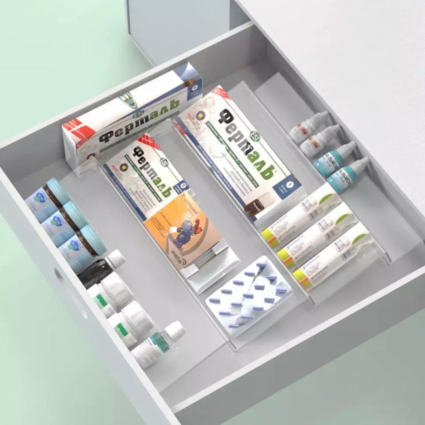 Geöffnete Schublade eines modernen, hellgrauen Schranks mit einem Acryl-Organizer, der verschiedene Medizinprodukte und Verbandsmaterialien übersichtlich hält. Die Schublade enthält gut sichtbar arrangierte Artikel wie Salben, Pflaster und Verpackungen, die mit pharmazeutischen Labels versehen sind. Der klare Organizer ermöglicht eine einfache Identifizierung und Zugänglichkeit der Gesundheitsprodukte.