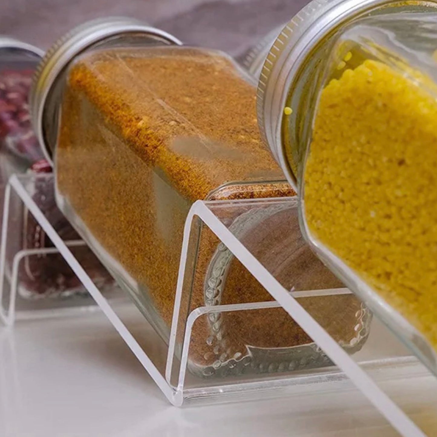 Nahaufnahme eines transparenten Acryl-Gewürzorganizers, der eine diagonale Anordnung verschiedener Gewürze in Glasbehältern zeigt. Links im Bild ist Kurkuma in einem umgedrehten Glas, gefolgt von einem weiteren Gewürz in einem aufrechten Glas, erkennbar an der gelben Farbe und den körnigen Texturen. Der Fokus auf die klaren Linien des Organizers unterstreicht seine praktische und ästhetische Eignung für moderne Küchen.