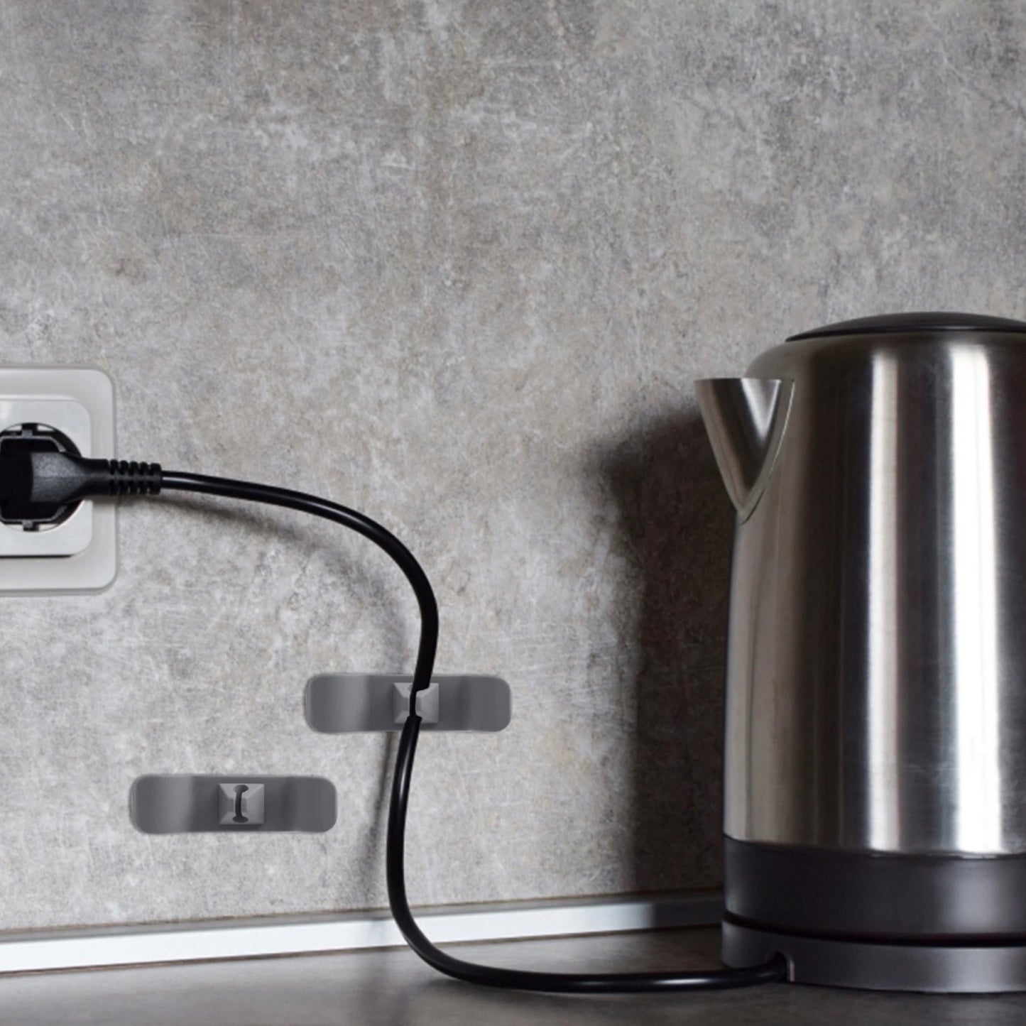Das Bild zeigt eine Küchenszene mit einem Edelstahl-Wasserkocher auf einer Arbeitsplatte und zwei grauen Kabelhaltern an der Wand, die das Kabel des Wasserkochers ordentlich führen. Die Kabelhalter sind strategisch platziert, um das Kabel sauber und aus dem Weg zu halten, während der Wasserkocher in Gebrauch ist.