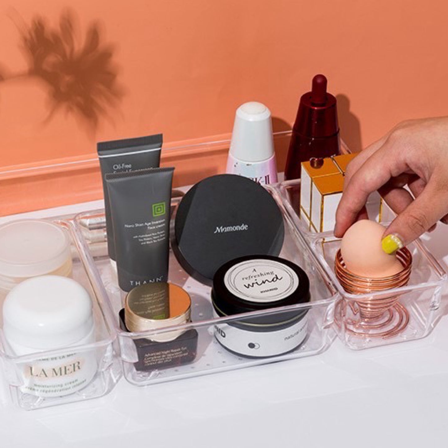 Ein transparenter Schubladen-Organizer, gefüllt mit verschiedenen hochwertigen Hautpflegeprodukten und Make-up-Artikeln. Zu sehen sind Cremes, ein Ölfreies Gesichtsgel, ein Puder, Lippenstift und ein Make-up-Schwamm, der gerade von einer Hand gehalten wird. Ideal für die übersichtliche und griffbereite Aufbewahrung von Kosmetikartikeln auf der Kommode oder im Badezimmer.
