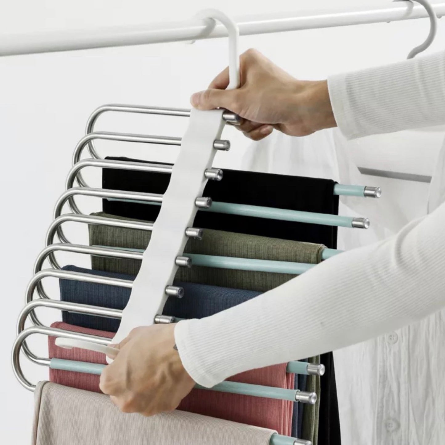 Eine Nahaufnahme einer Hand, die einen innovativen, mehrstufigen Kleiderbügel in einem Kleiderschrank anpasst. Die Kleiderbügel bieten mehrere Reihen zur effizienten Aufbewahrung von Hosen und anderen Kleidungsstücken in verschiedenen Farben. Diese Lösung ist ideal für die optimale Nutzung von begrenztem Schrankraum und hält Kleidung organisiert und leicht zugänglich.