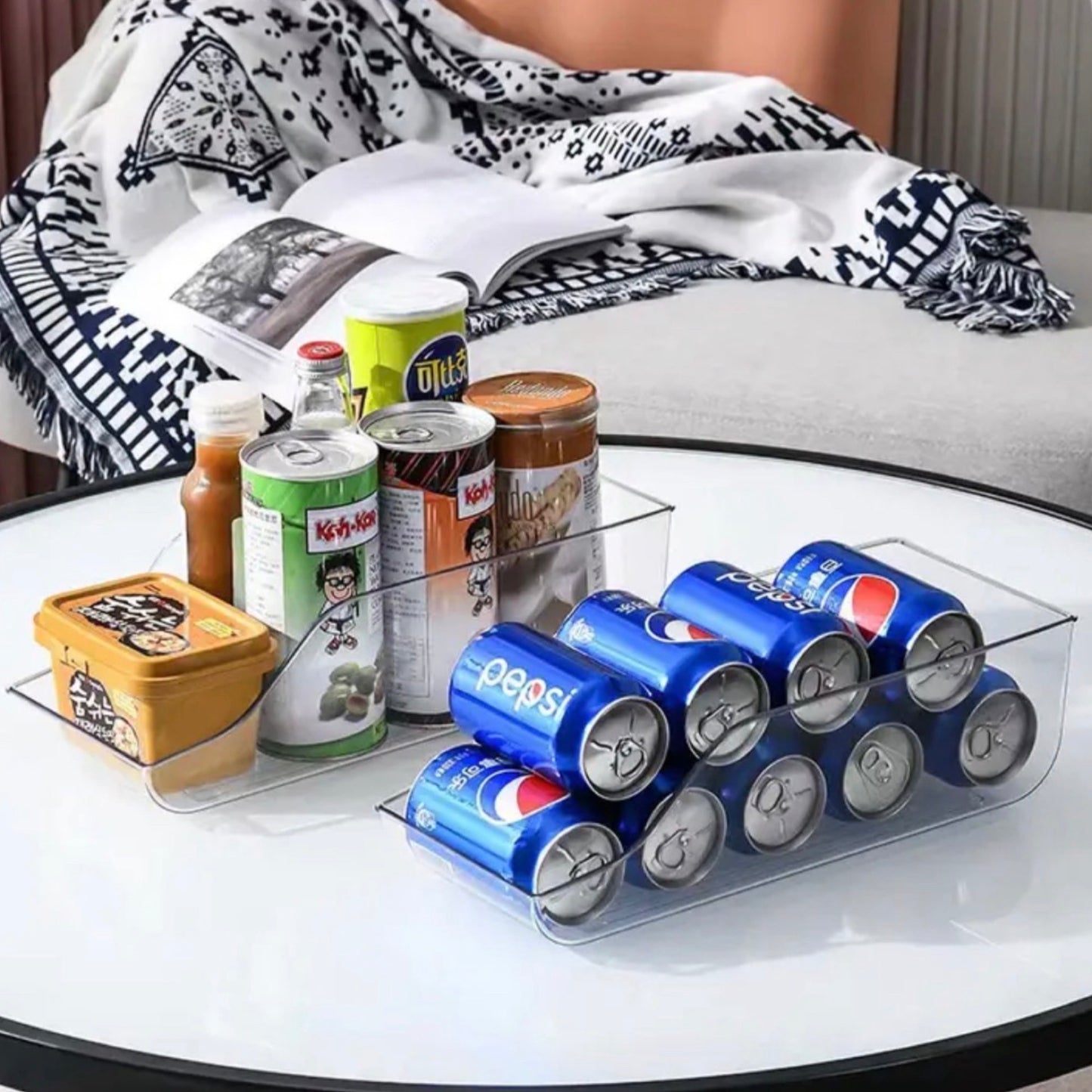 Ein transparenter Dosenhalter auf einem weißen runden Tisch, bestückt mit blauen Pepsi-Dosen, neben verschiedenen Lebensmitteln und einem offenen Buch, im Hintergrund einer schwarz-weißen Decke.