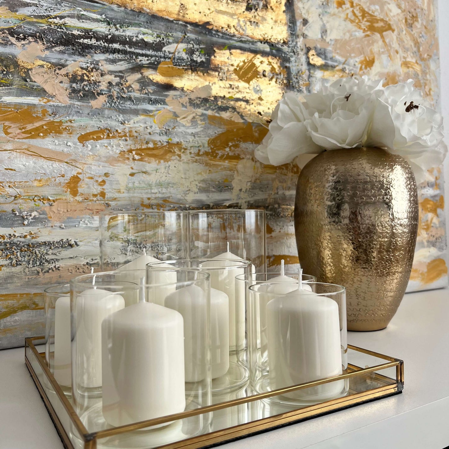 Windlichter mit weißen Kerzen auf einem goldgerahmten Spiegeltablett, ergänzt durch eine dekorative goldene Vase mit weißer Blume vor strukturierter Wand – stilvolle Tischdekoration im modernen Look.