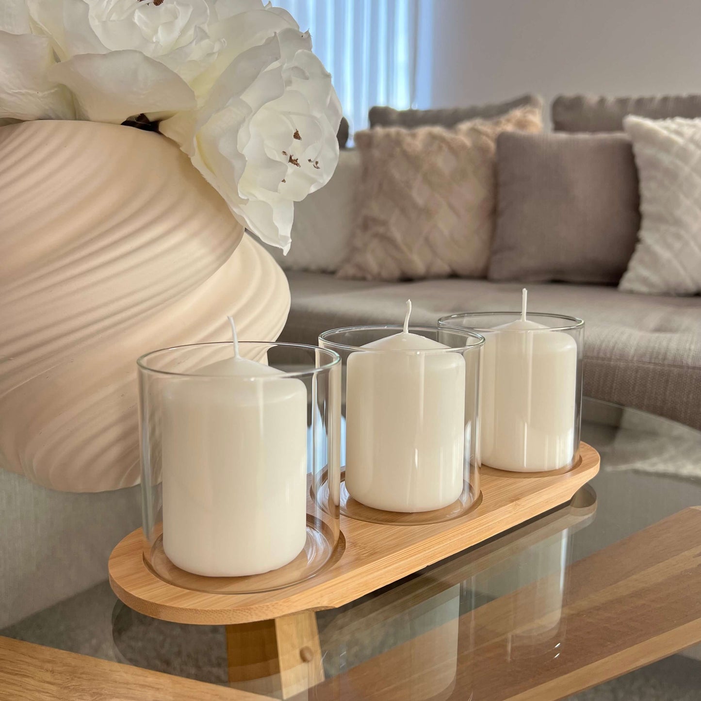 Drei kleine Windlichter mit weißen Kerzen auf einem Holztablett,Keramikvase in Creme mit weißen Kunstblumen im Hintergrund ein Sofa mit strukturierten Kissen.