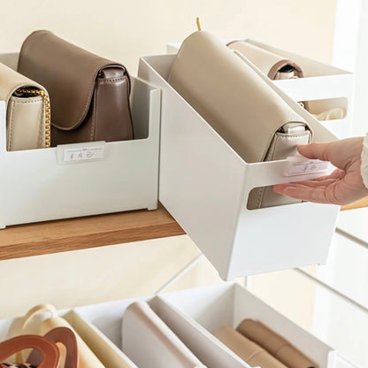 Stapelbare Aufbewahrungsbox mit Griff, gezeigt mit verschiedenen eleganten Handtaschen im Inneren. Die weißen Boxen sind auf einem Holzregal arrangiert und bieten eine ansprechende und ordentliche Lagermöglichkeit für Accessoires. Eine Hand zieht an einem Griff, um eine der Schubladen zu öffnen, in der eine beige Handtasche sichtbar ist, was die einfache Handhabung demonstriert.