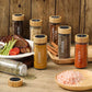 Eine rustikale Tischszene mit einer Auswahl an Gewürzstreuern, etikettiert mit ‘Curry’, ‘Pfeffer’, ‘Cajun’ und anderen. Sie sind auf einem Holztisch platziert neben einem Teller mit einem saftigen Steak, Spargel und Tomaten, was die Verwendung der Gewürze in der Küche zum Kochen und Servieren von Mahlzeiten unterstreicht.