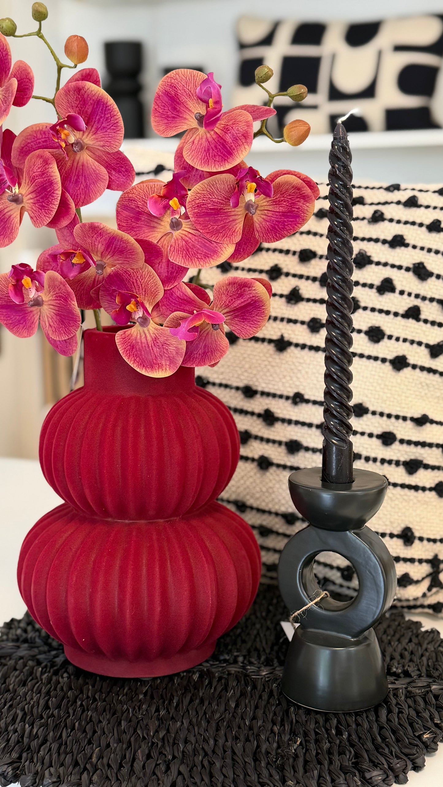 schöne arngierte rote Vase mit rote Orchidee,neben hat schwarze Kerzenständer mit schwarze Kerze.Hinten die Vase hat weiss-schwarze Kissen
