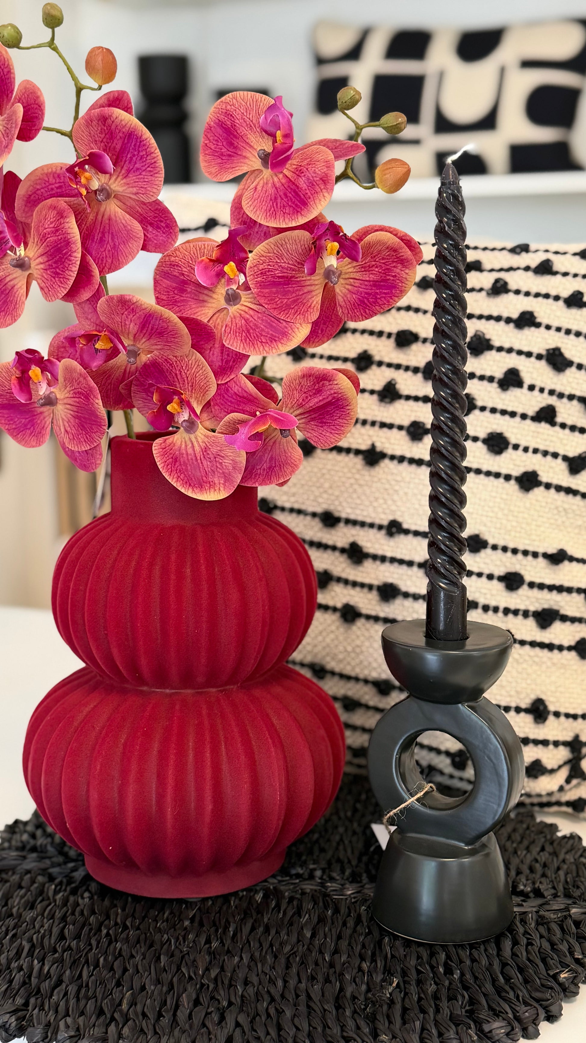 schöne arngierte rote Vase mit rote Orchidee,neben hat schwarze Kerzenständer mit schwarze Kerze.Hinten die Vase hat weiss-schwarze Kissen