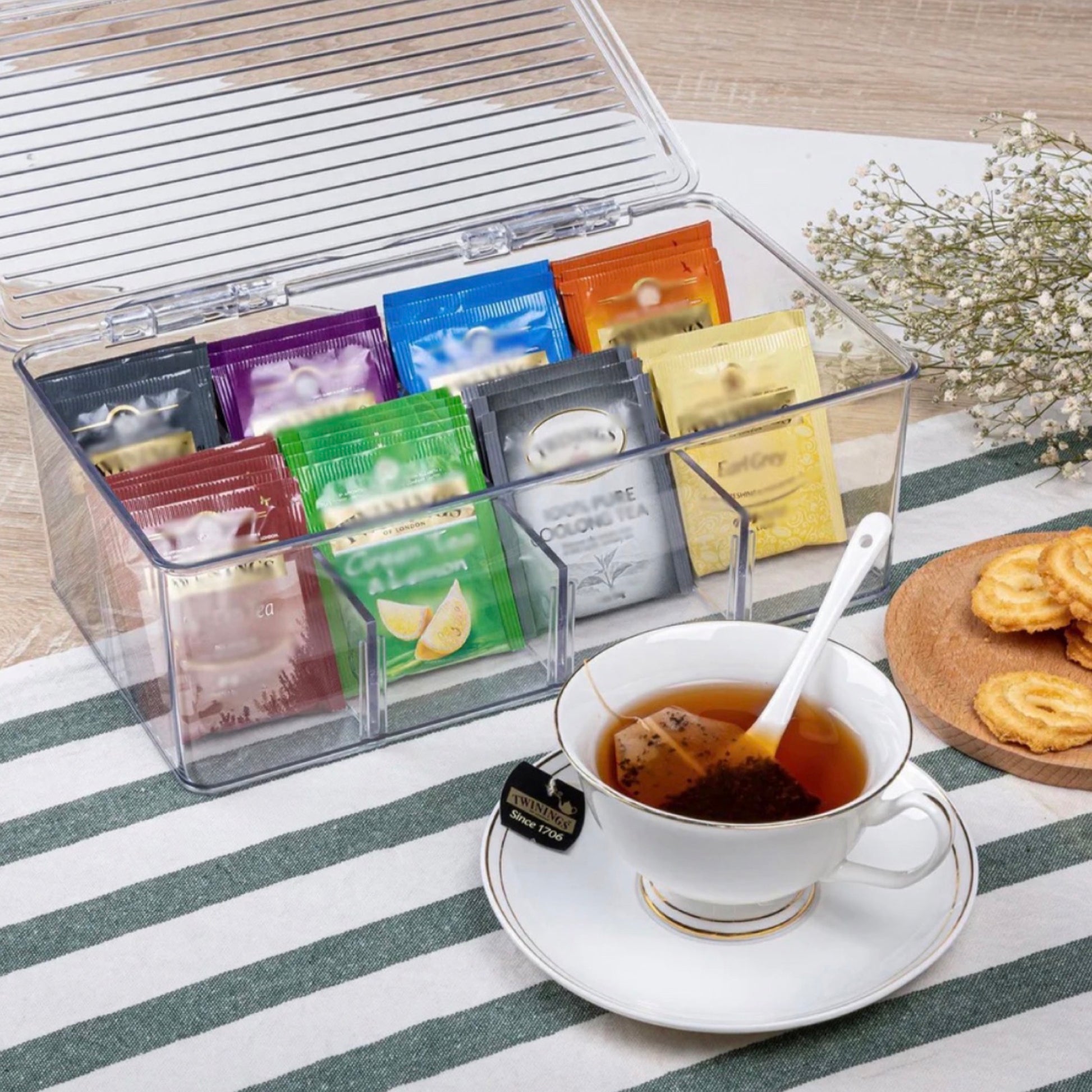 Ein gemütlicher Teemoment mit einer transparenten Teekiste voller bunter Teebeutel-Variationen geöffnet auf einem Tisch. Neben der Box ist eine elegante Teetasse auf einer Untertasse, gefüllt mit heißem Tee, und auf einem Teller daneben liegen zwei Kekse, arrangiert auf einem gestreiften Tischtuch.