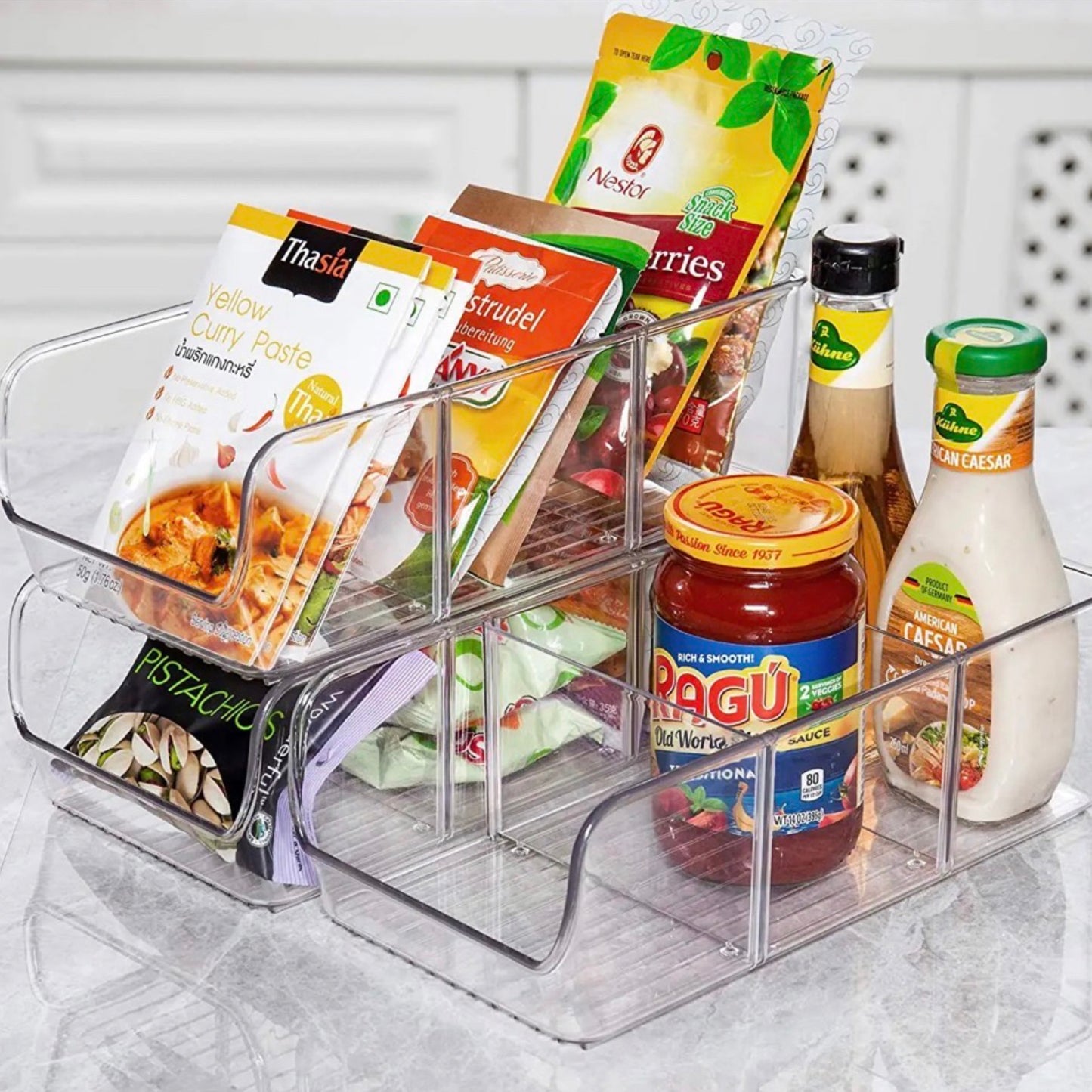 Transparenter iDesign Acryl-Aufbewahrungsbehälter auf einer Küchenarbeitsplatte, gefüllt mit einer Vielfalt an Lebensmitteln. Zu sehen sind Pakete mit Gelber Currypaste, Apfelstrudel-Mix, getrockneten Beeren und Pistazien, sowie Gläser mit Tomatensauce und Salatdressing. Der Organizer sortiert die Artikel übersichtlich und macht sie leicht zugänglich.