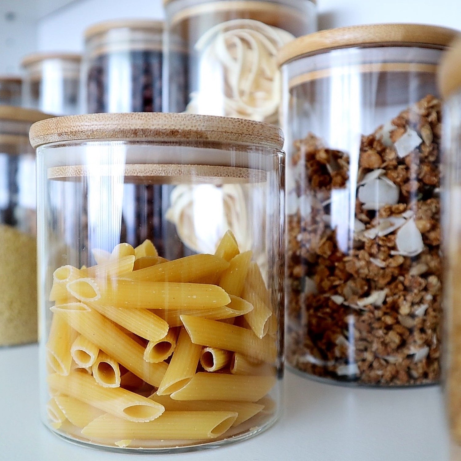 Vorratsglas mit Bambusdeckel, gefüllt mit Penne-Nudeln. Im Hintergrund weitere Gläser mit Pasta und Müsli – ideal für stilvolle Küchenorganisation
