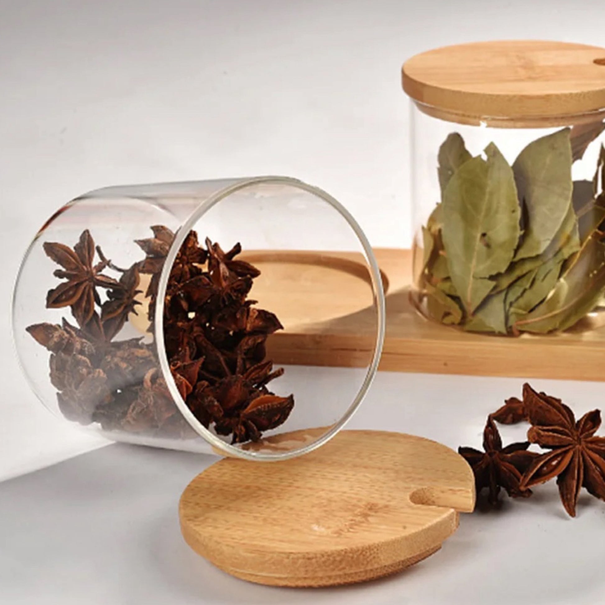 Ein umgekipptes Gewürzglas zeigt seine offene Seite mit verstreuten Sternanis auf einer hellen Oberfläche. Im Hintergrund steht ein gefülltes Glas mit Lorbeerblättern auf einem eleganten Bambustablett, neben einem aufrecht stehenden Bambusdeckel mit einer kreisförmigen Griffaussparung.