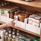 In einer Küchenablage unterstützen weiße, stapelbare Aufbewahrungsboxen die strukturierte Lagerung von verschiedenen Lebensmitteln. Eine Hand deutet auf die Boxen, die Joghurts, Kaffeegetränke und Desserts enthalten. Unter dem Regal sind weitere Getränke und Snacks zu sehen, alles ist leicht zugänglich und ordentlich angeordnet.