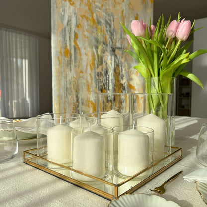 Windlichter aus Glas mit weißen Kerzen auf einem goldgerahmten Tablett, dekoriert mit rosa Tulpen in einer Vase und einem abstrakten Wandbild im Hintergrund