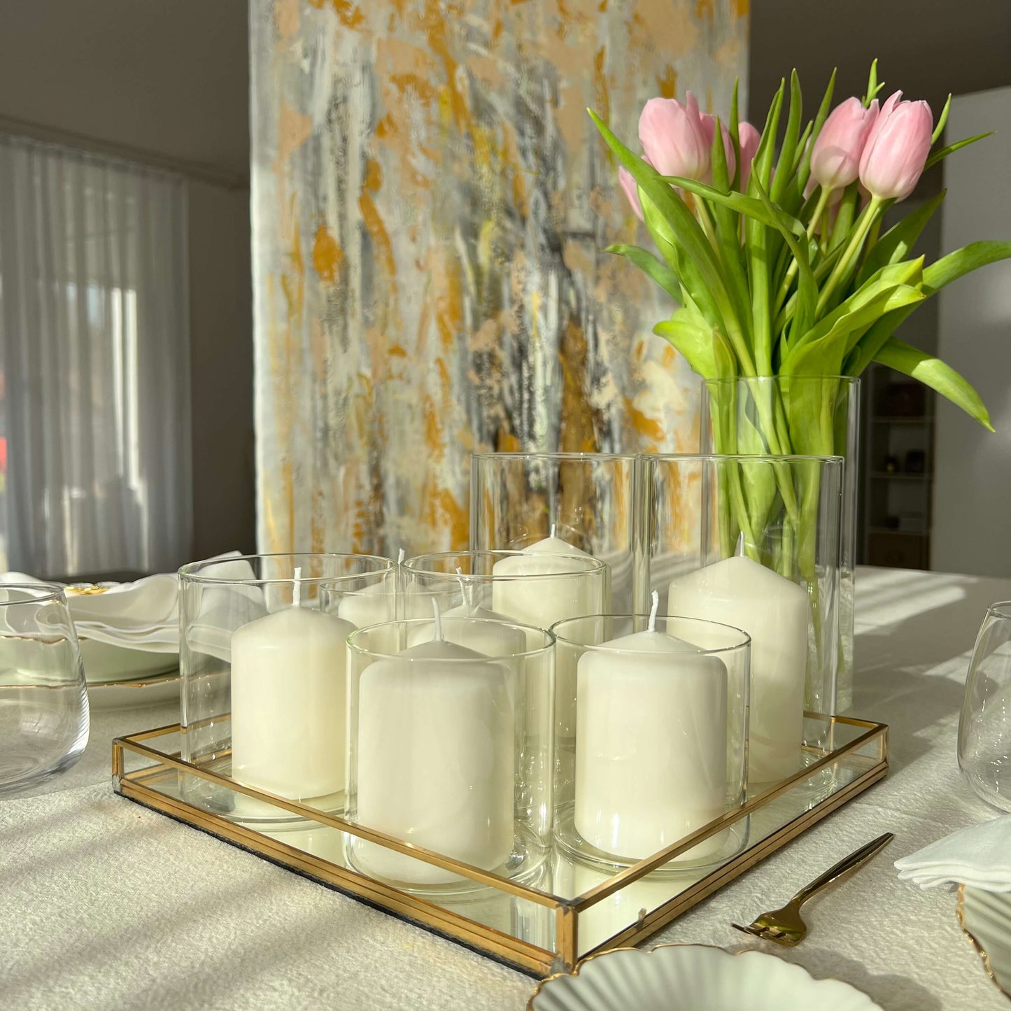 Windlichter aus Glas mit weißen Kerzen auf einem goldgerahmten Tablett, dekoriert mit rosa Tulpen in einer Vase und einem abstrakten Wandbild im Hintergrund