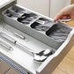 Besteck-Organizer mit fünf Fächern in einer Schublade, gefüllt mit glänzendem Edelstahlbesteck. Eine Hand entnimmt ein Besteckstück. Unterhalb des Organizers liegen ein Schneebesen und zwei große Servierlöffel auf einer weißen Schubladenfläche.
