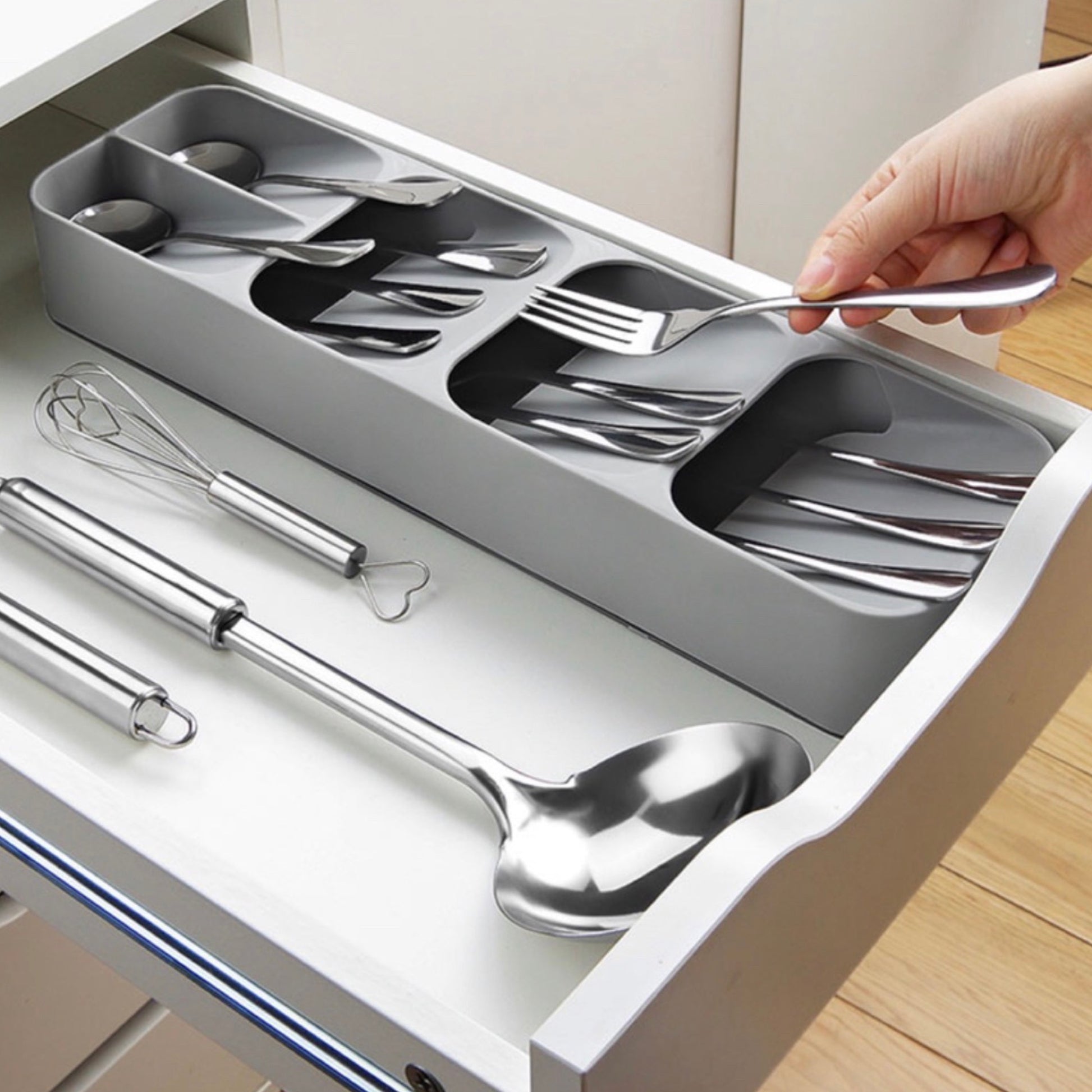 Besteck-Organizer mit fünf Fächern in einer Schublade, gefüllt mit glänzendem Edelstahlbesteck. Eine Hand entnimmt ein Besteckstück. Unterhalb des Organizers liegen ein Schneebesen und zwei große Servierlöffel auf einer weißen Schubladenfläche.