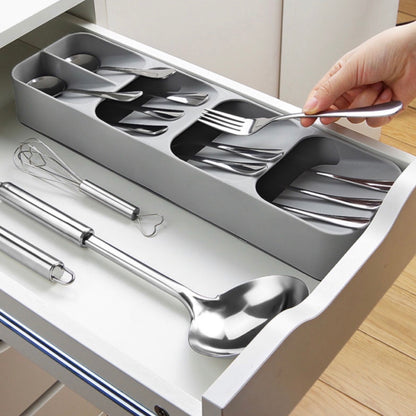 Besteck-Organizer mit fünf Fächern in einer Schublade, gefüllt mit glänzendem Edelstahlbesteck. Eine Hand entnimmt ein Besteckstück. Unterhalb des Organizers liegen ein Schneebesen und zwei große Servierlöffel auf einer weißen Schubladenfläche.
