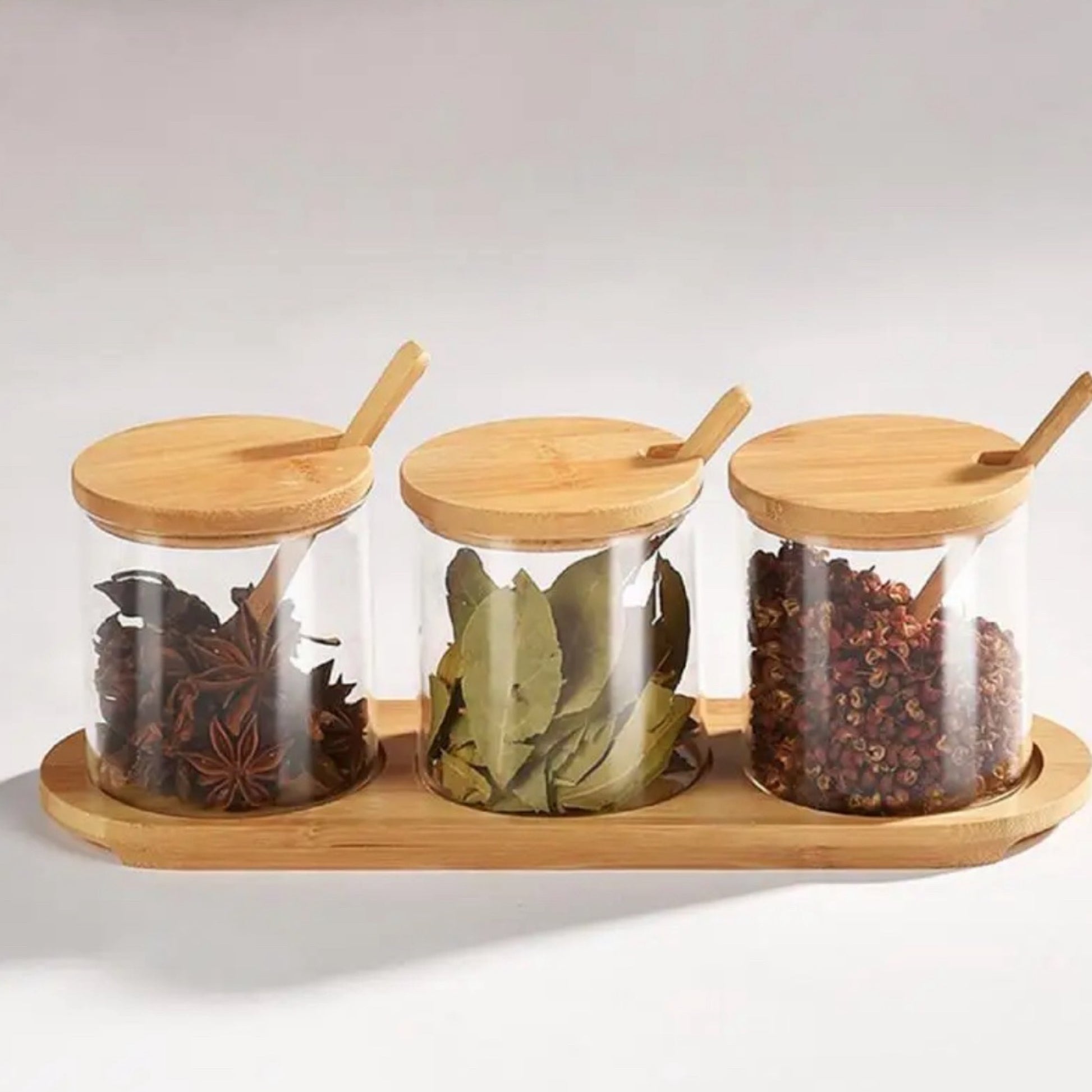 Drei klare Gewürzgläser mit eleganten, passenden Bambusdeckeln auf einem geschwungenen Bambustablett. Jedes Glas enthält ein anderes Gewürz: Sternanis, Lorbeerblätter und Szechuanpfeffer, jeweils sichtbar und reich an Textur. Inklusive sind kleine Bambuslöffel, die auf den Gläsern liegen, bereit, die aromatischen Inhalte zu entnehmen.