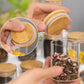 Eine Hand hält ein transparentes Gewürzglas, gefüllt mit einer bunten Gewürzmischung, und einen Bambusdeckel mit Silikonband, der das Öffnen und Schließen erleichtert. Im Hintergrund sind weitere gefüllte Gläser unscharf zu sehen, was die Nutzungsvielfalt hervorhebt.