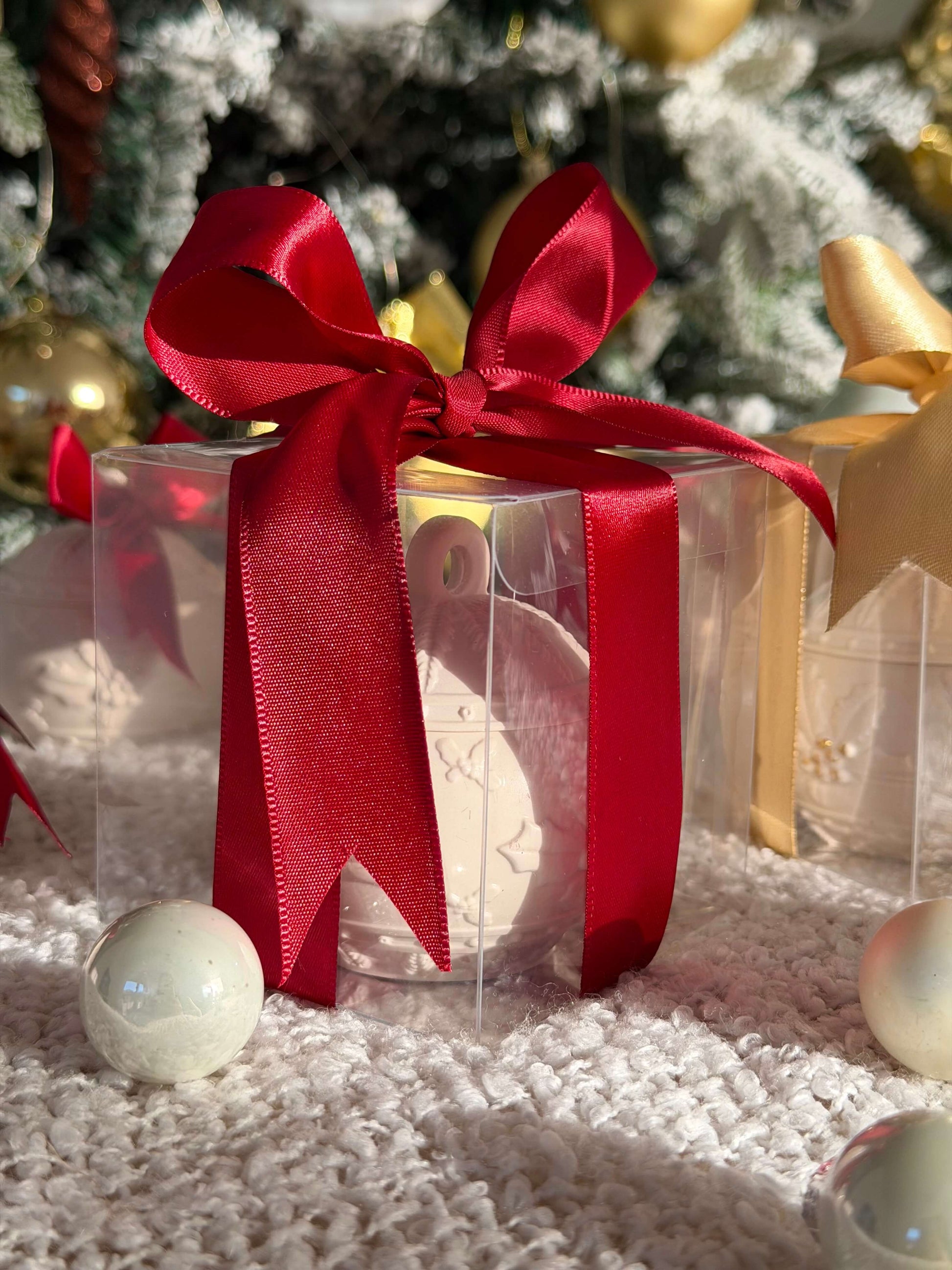 Handgefertigte Weihnachtskugel in transparenter Geschenkbox mit großer roter Schleife, dekorativ vor einem festlich geschmückten Weihnachtsbaum arrangiert