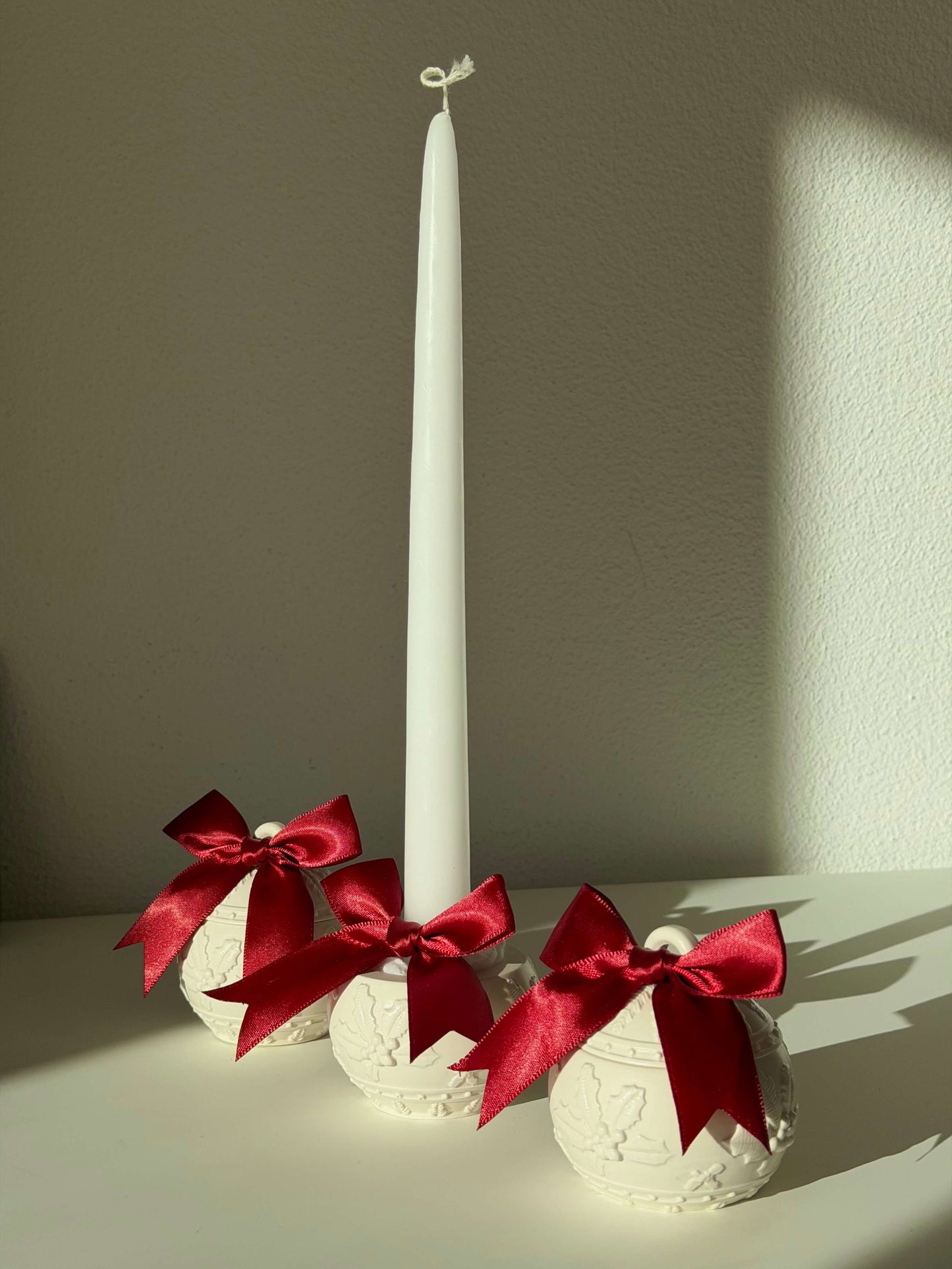 Drei handgefertigte Weihnachtskugel-Kerzenhalter mit roten Schleifen, dekorativ neben einer weißen Stabkerze im warmen Sonnenlicht arrangiert