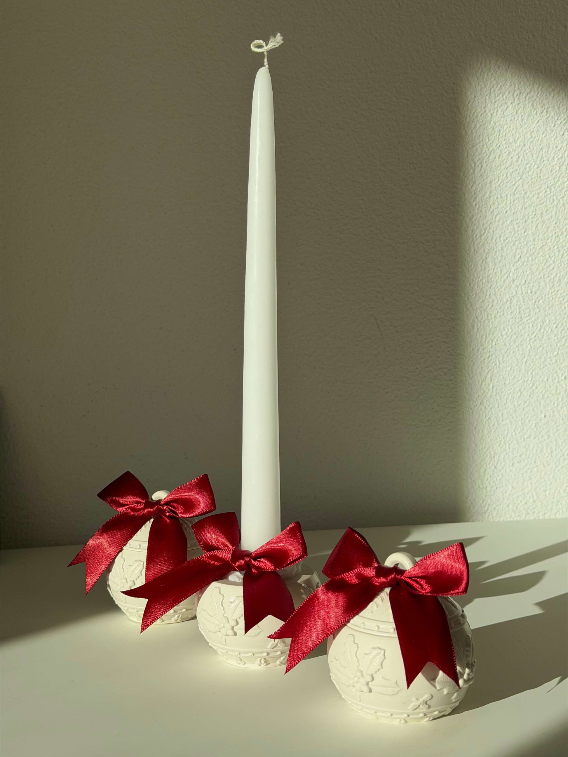 Drei handgefertigte Weihnachtskugel-Kerzenhalter mit roten Schleifen, dekorativ neben einer weißen Stabkerze im warmen Sonnenlicht arrangiert