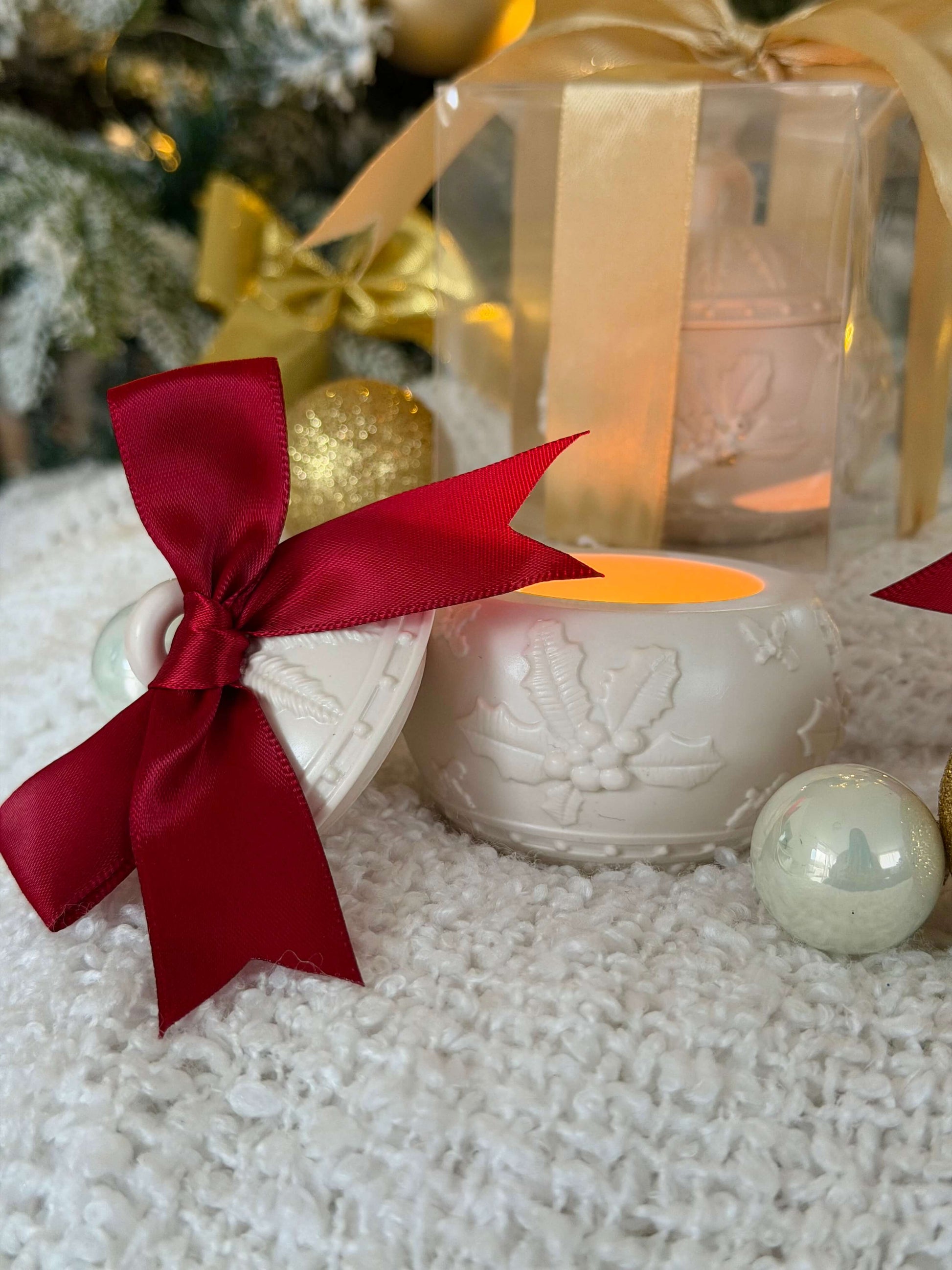 Geöffneter handgefertigter Weihnachtskugel-Kerzenhalter mit roter Schleife und warmem Licht, dekorativ vor einer Geschenkbox mit goldener Schleife arrangiert