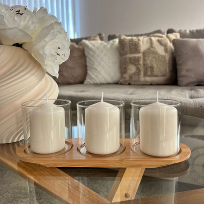 Keramikvase in Creme mit weiße blumen, elegante drei kleine Windlichter mit weißen Kerzen auf einem Holztablett, im Hintergrund ein Sofa mit crem Kissen.