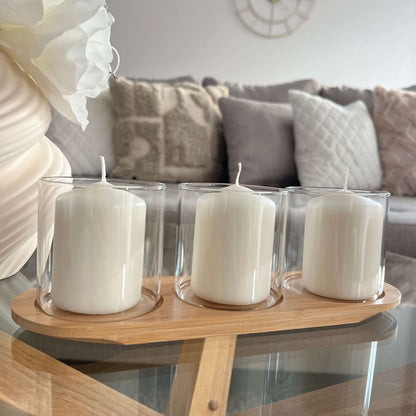 Drei Windlichtgläser mit weißen Kerzen auf einem ovalen Holztablett, arrangiert auf einem Glastisch vor einem gemütlichen Sofa mit Kissen.