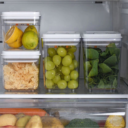 Vier stapelbare Vorratsdosen mit durchsichtigen Wänden und weißen Deckeln im Kühlschrank, befüllt mit frischen Lebensmitteln zur Schau stellend. Die linke Dose enthält Sauerkraut, die mittlere grüne Weintrauben und die rechte frische grüne Blätter, vermutlich Kräuter. Darunter im Kühlschrank sind weitere Gemüsesorten zu sehen, was auf eine gesunde und organisierte Lagerung hindeutet.