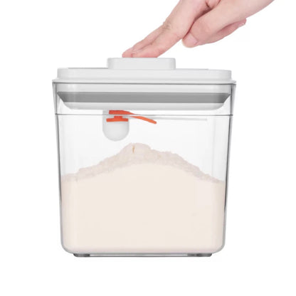 Eine Hand bedient den Pop-up-Verschluss einer transparenten Vorratsdose, die teilweise mit Mehl gefüllt ist. Der orange Verschlussmechanismus an der Frontseite, mit dem man einen Löffel oder eine Messschaufel anhängen kann, ist deutlich sichtbar, was die praktische Funktionalität der Dose unterstreicht.