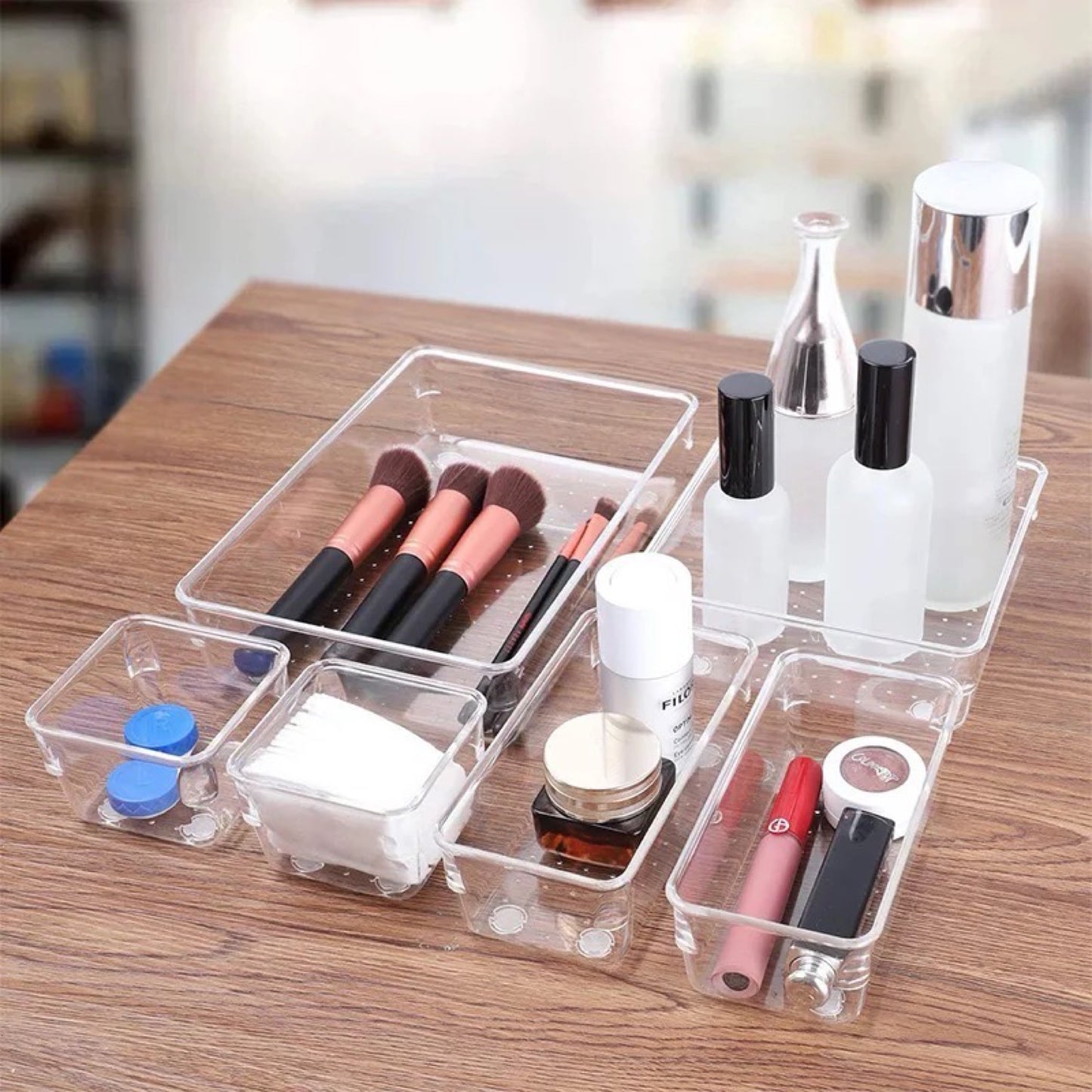 Ein transparenter Kosmetik-Organizer auf einem Holztisch, gefüllt mit Schönheitsprodukten. Er enthält vier separate Fächer mit Makeup-Pinseln, Gesichtsreinigungsflaschen, Wattepads, einer Gesichtscreme, Augenpflegeprodukten und Lippenstiften. Dieser Organizer bietet eine effiziente und stilvolle Lösung, um Kosmetikartikel geordnet und leicht zugänglich zu halten.