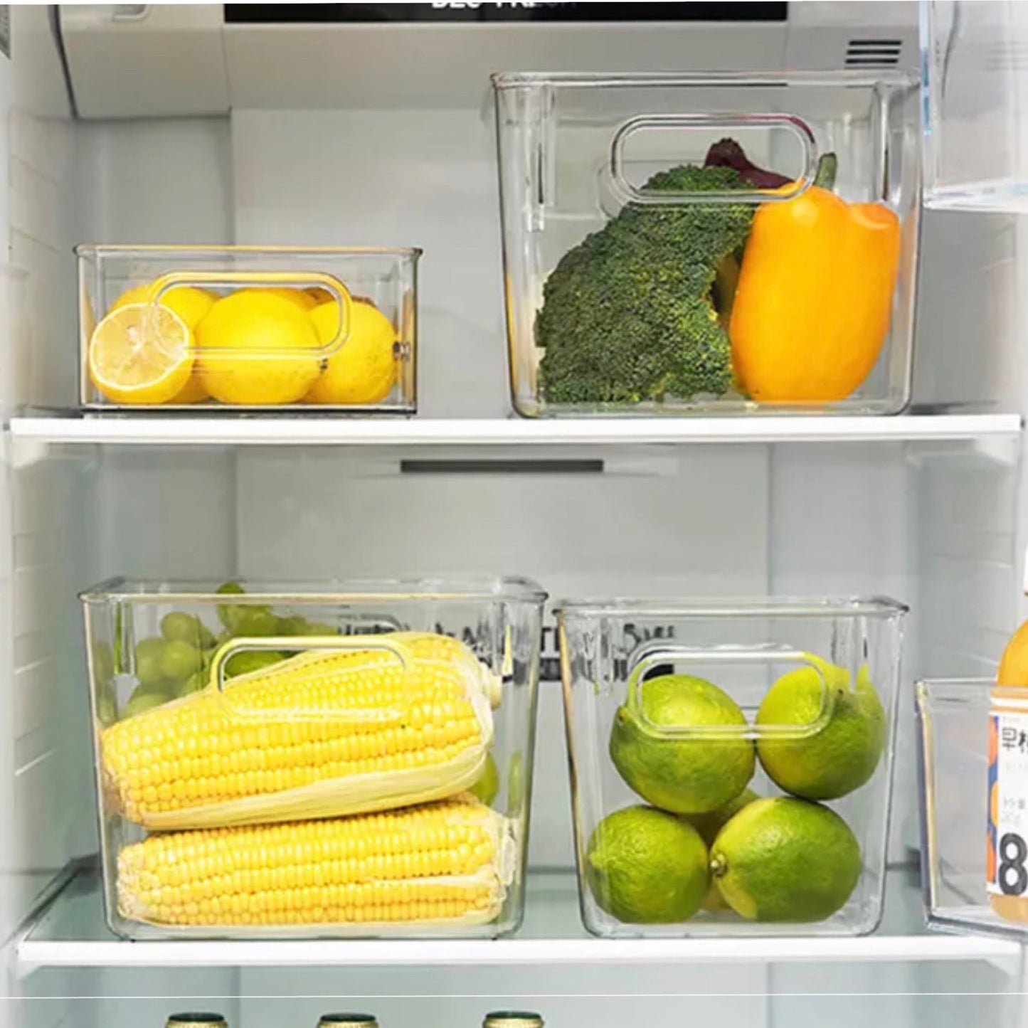 Transparente Aufbewahrungsboxen im Kühlschrank mit verschiedenen Lebensmitteln. Oben links ist eine Box mit Zitronenscheiben und ganzen Zitronen, daneben eine mit Brokkoli und einer gelben Paprika. Unten links befindet sich eine Box mit frischen Maiskolben und rechts daneben eine mit grünen Limetten. Die Anordnung zeigt eine saubere und organisierte Lagerung.