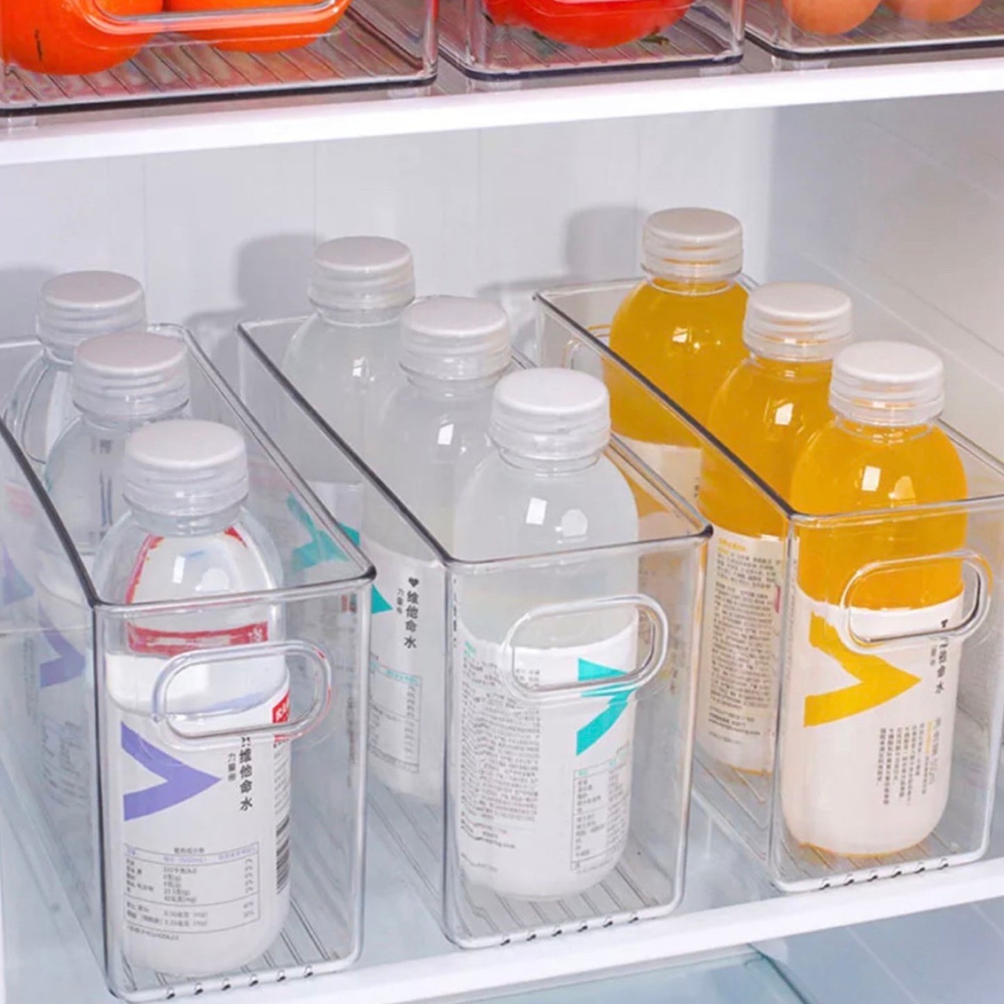 Transparente Aufbewahrungsboxen im Kühlschrank, befüllt mit ordentlich ausgerichteten Wasserflaschen und farbigen Getränken. Die Etiketten der Flaschen sind in verschiedenen Sprachen beschriftet, was auf ein multikulturelles Sortiment hindeutet. Die Boxen ermöglichen eine klare Sichtbarkeit und leichten Zugang zu den Getränken und nutzen den verfügbaren Platz effizient.