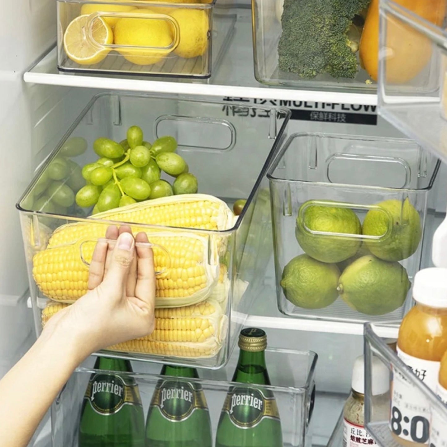 Transparente Aufbewahrungsboxen im Kühlschrank, eine mit frischen Maiskolben, eine mit grünen Trauben und eine dritte mit Limetten. Eine Hand greift nach einer der Maiskolben, was die praktische Zugänglichkeit der Boxen hervorhebt. Unten im Kühlschrank sind Getränkeflaschen zu sehen. Diese Organisation hilft dabei, Lebensmittel frisch und übersichtlich zu halten.